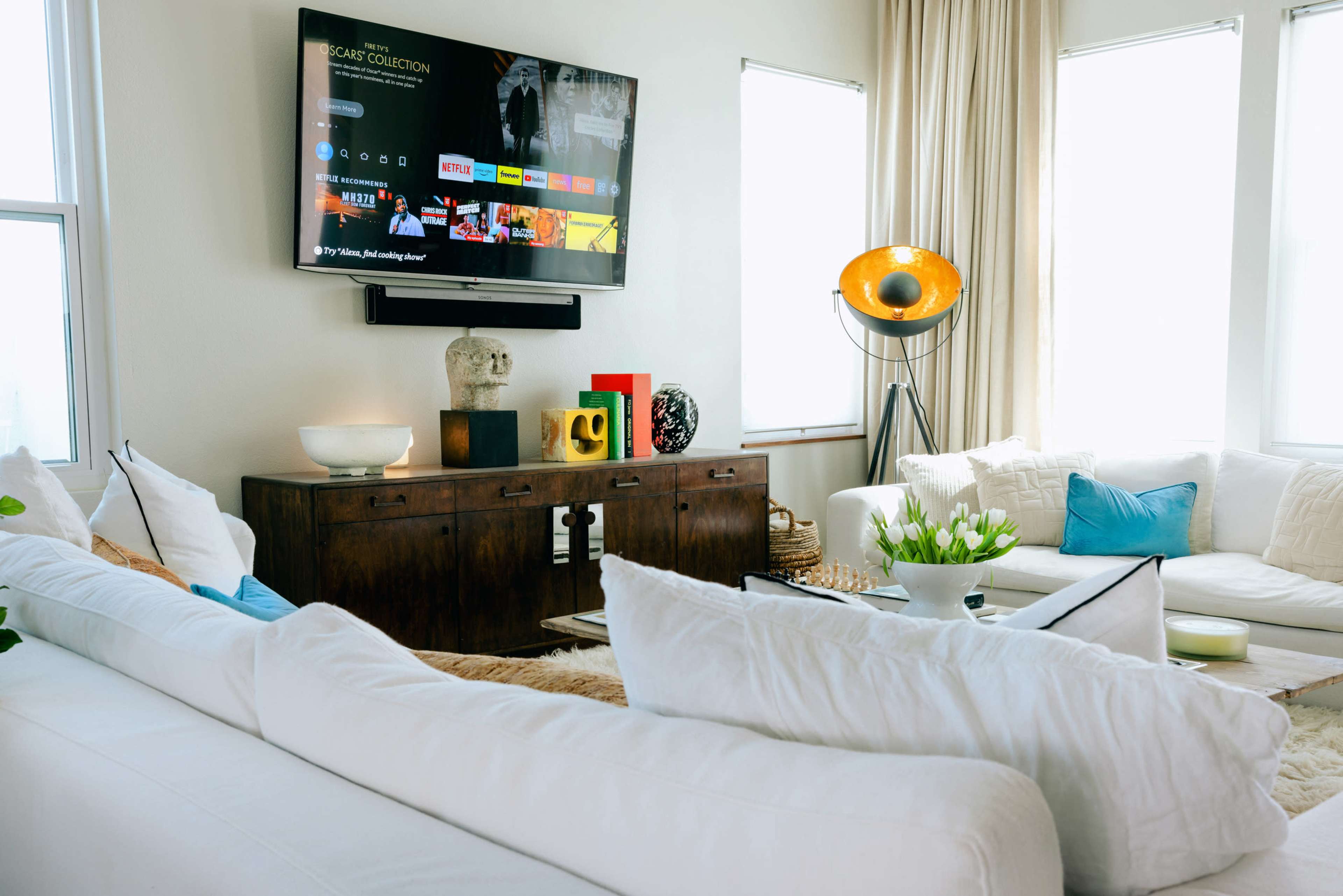 A modern living room features a large TV mounted on the wall, a wooden console table, and seating arrangements with light-colored sofas and decorative pillows.