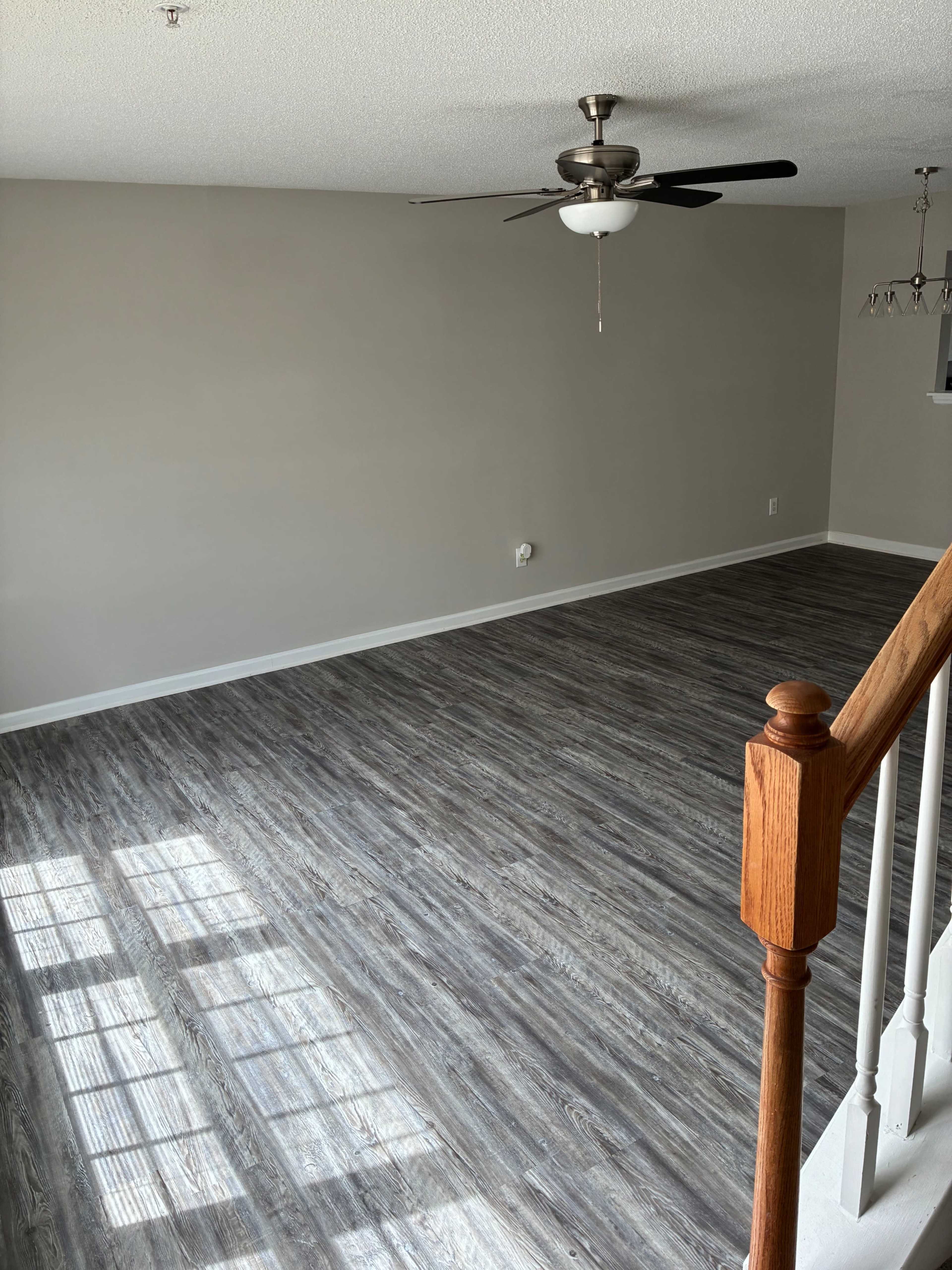 The image shows an empty room with gray walls, a ceiling fan, and new wooden flooring, illuminated by natural light.