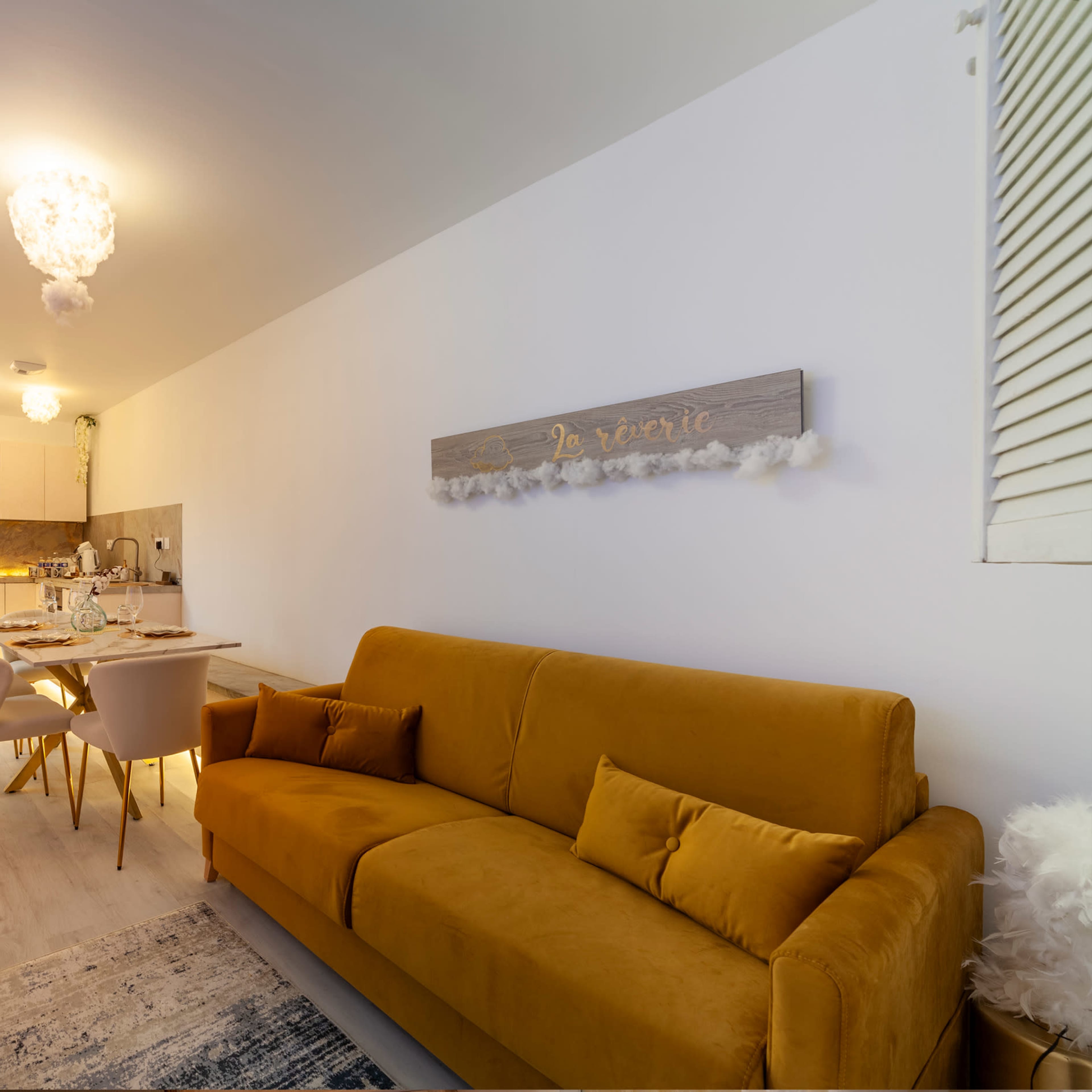 The image shows a cozy living space featuring a mustard-colored sofa, a dining area with white chairs, and a decorative wall sign.