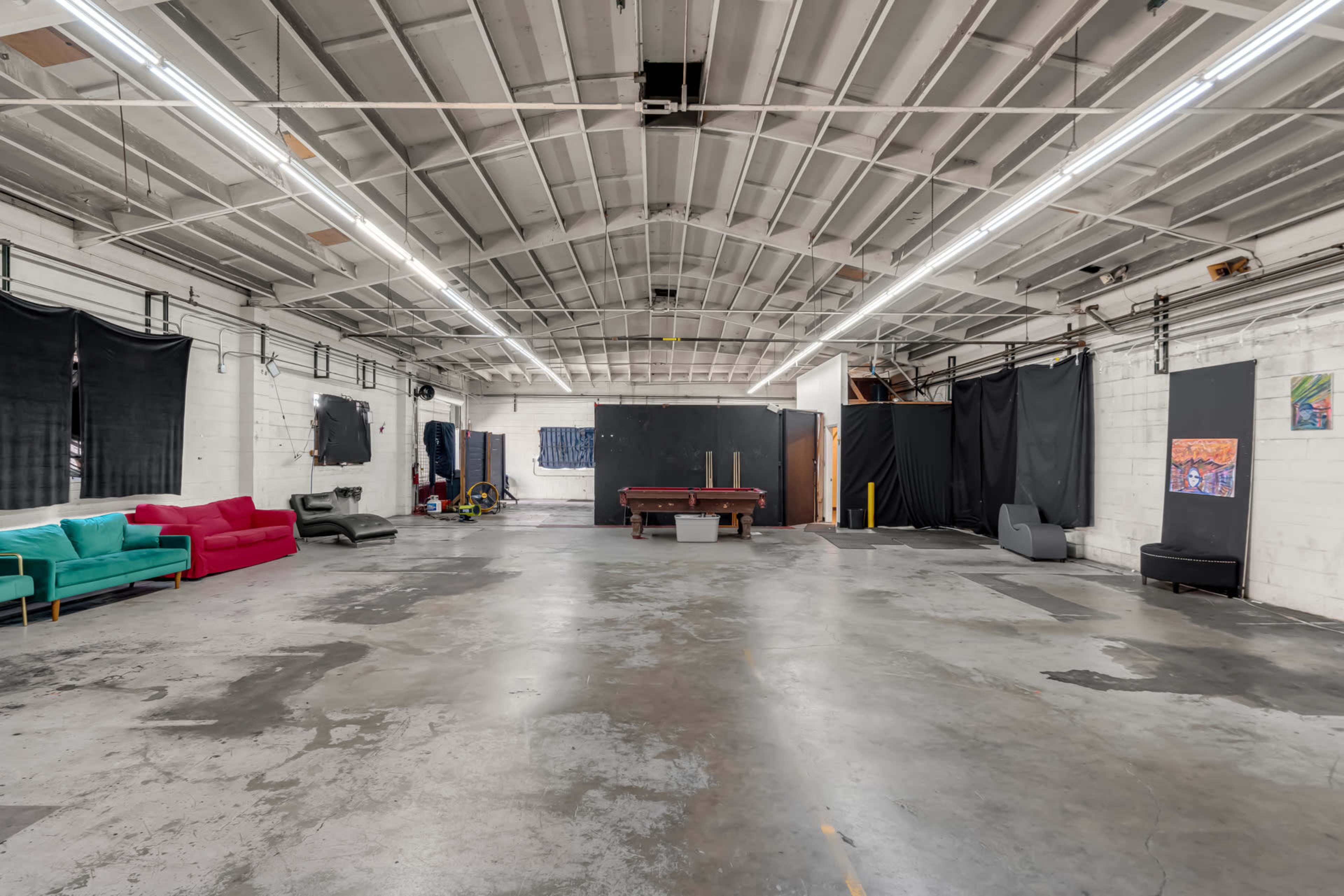 The image shows a spacious warehouse interior with high ceilings, bare concrete floors, and a pool table in the center, flanked by colorful sofas and black curtains along the walls.