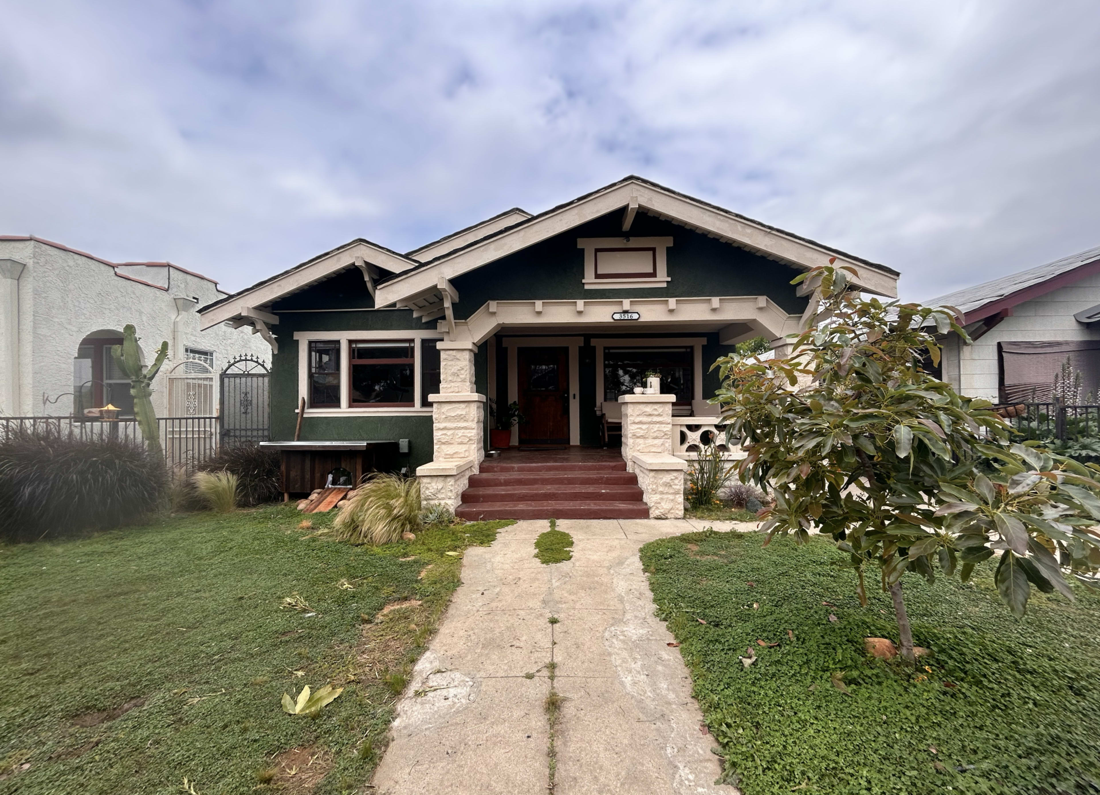The image shows a green bungalow-style house with a porch, surrounded by grass and a small tree in the front yard.