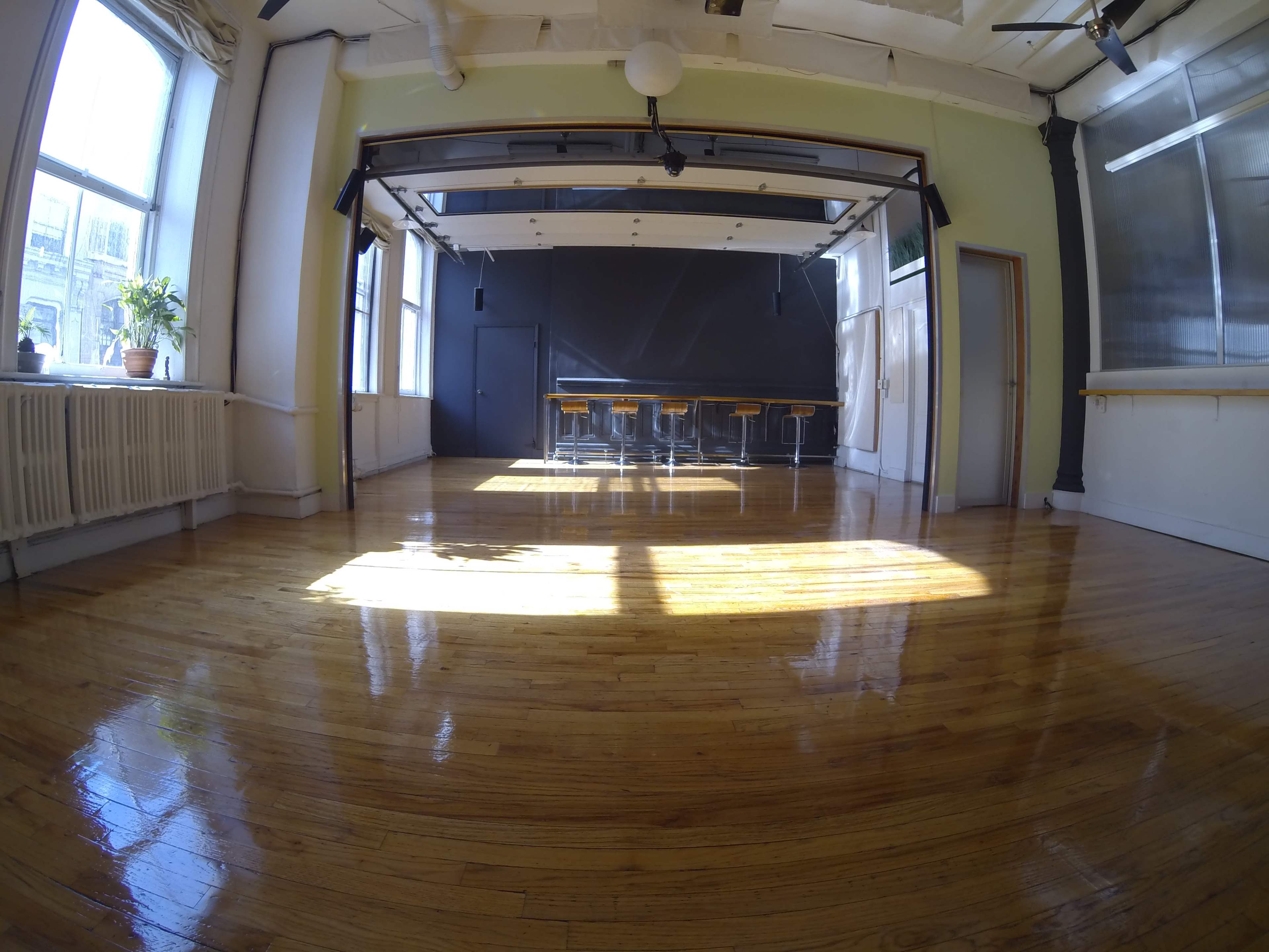 The image shows a spacious, bright room with polished wooden floors, large windows, and a table with several stools against a dark wall.