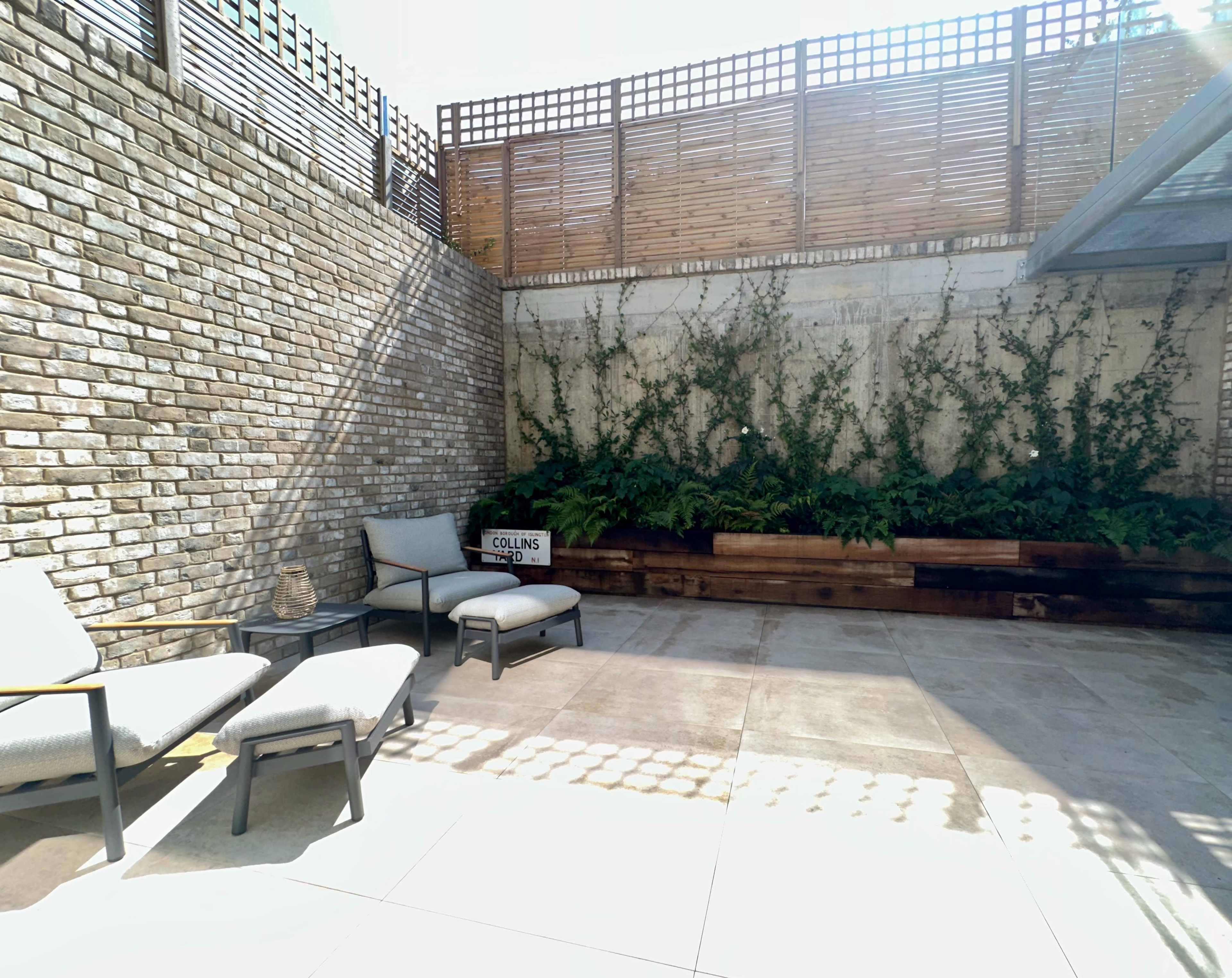 The image shows a minimalist outdoor patio area featuring two lounge chairs, a small side table, and a vertical garden against a brick wall.