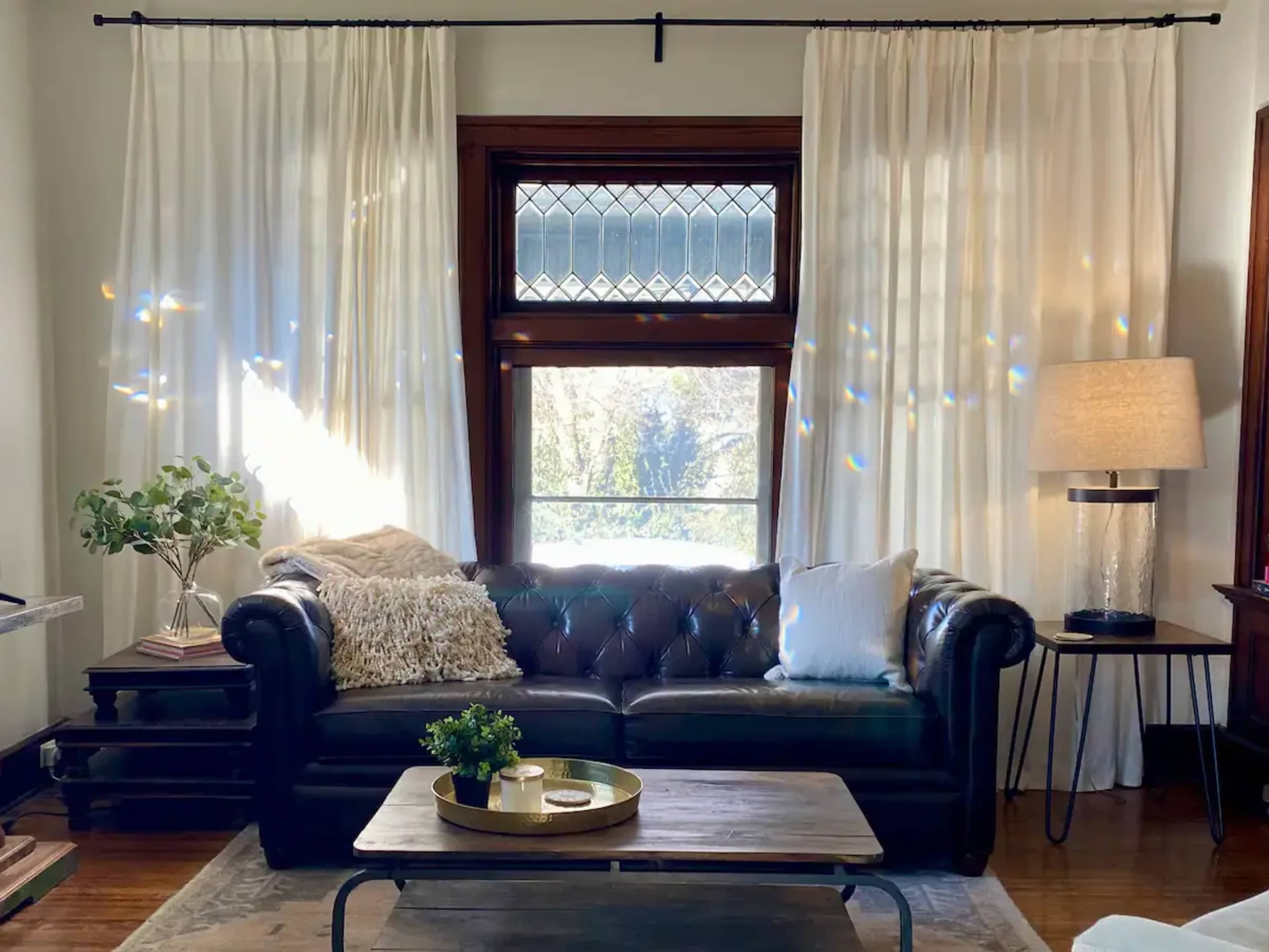 A leather sofa is positioned in a living room, accompanied by a coffee table and a floor lamp, with sheer curtains framing a window that lets in natural light.