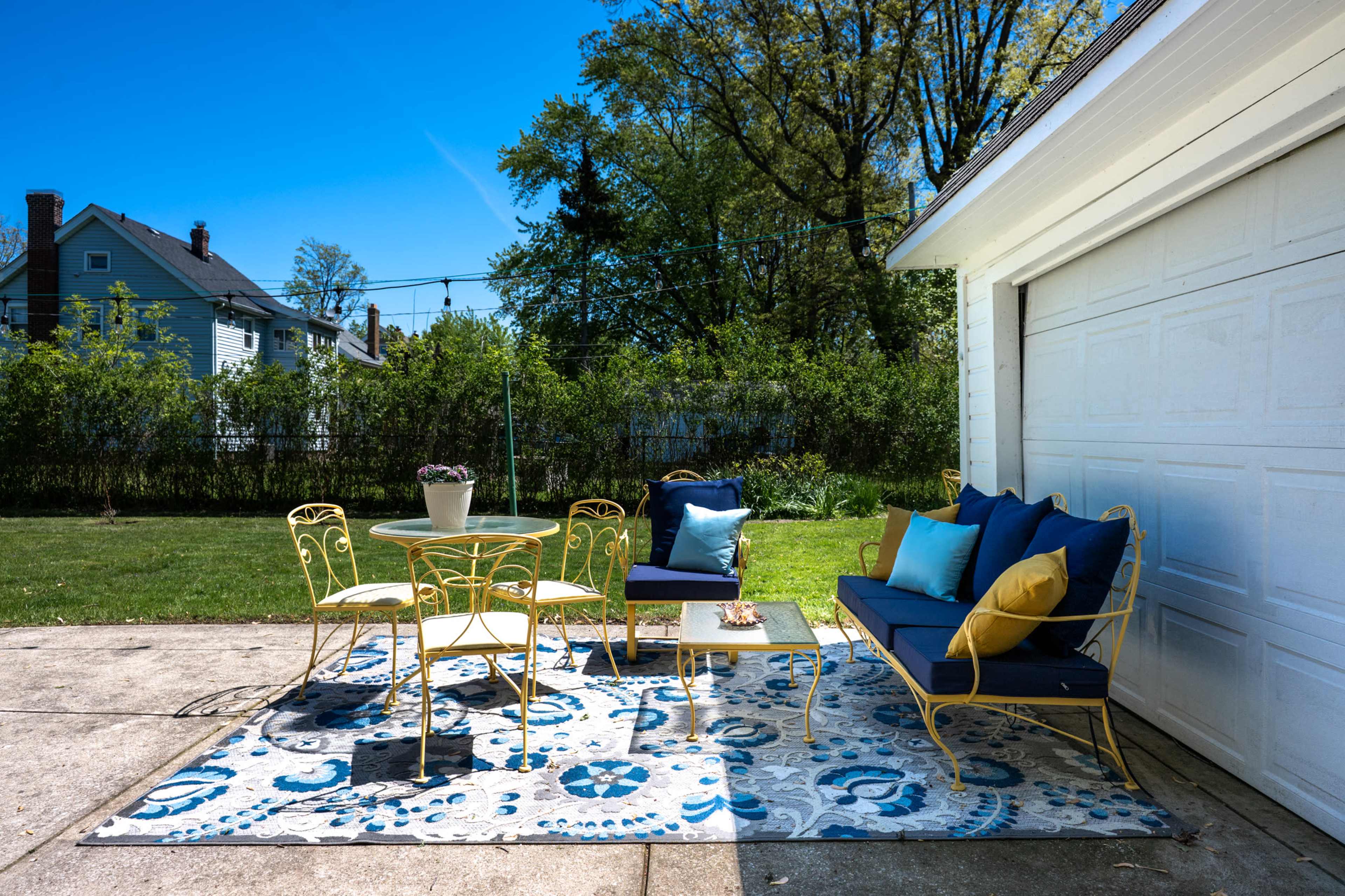 A patio area features a round table with chairs, a sofa, and a large area rug, set against a backdrop of greenery and a clear blue sky.