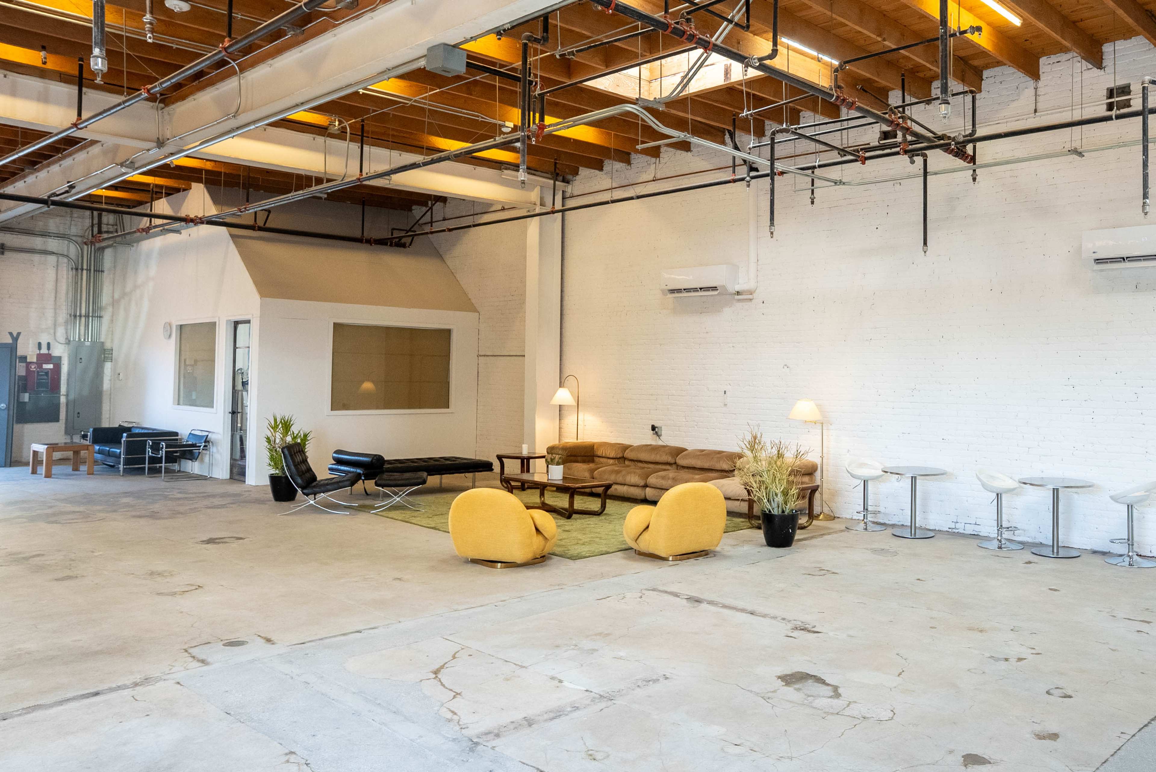The image shows a spacious, open-concept interior with a seating area featuring a brown couch, a black lounge chair, two yellow armchairs, and a small round table, all placed on a green rug.