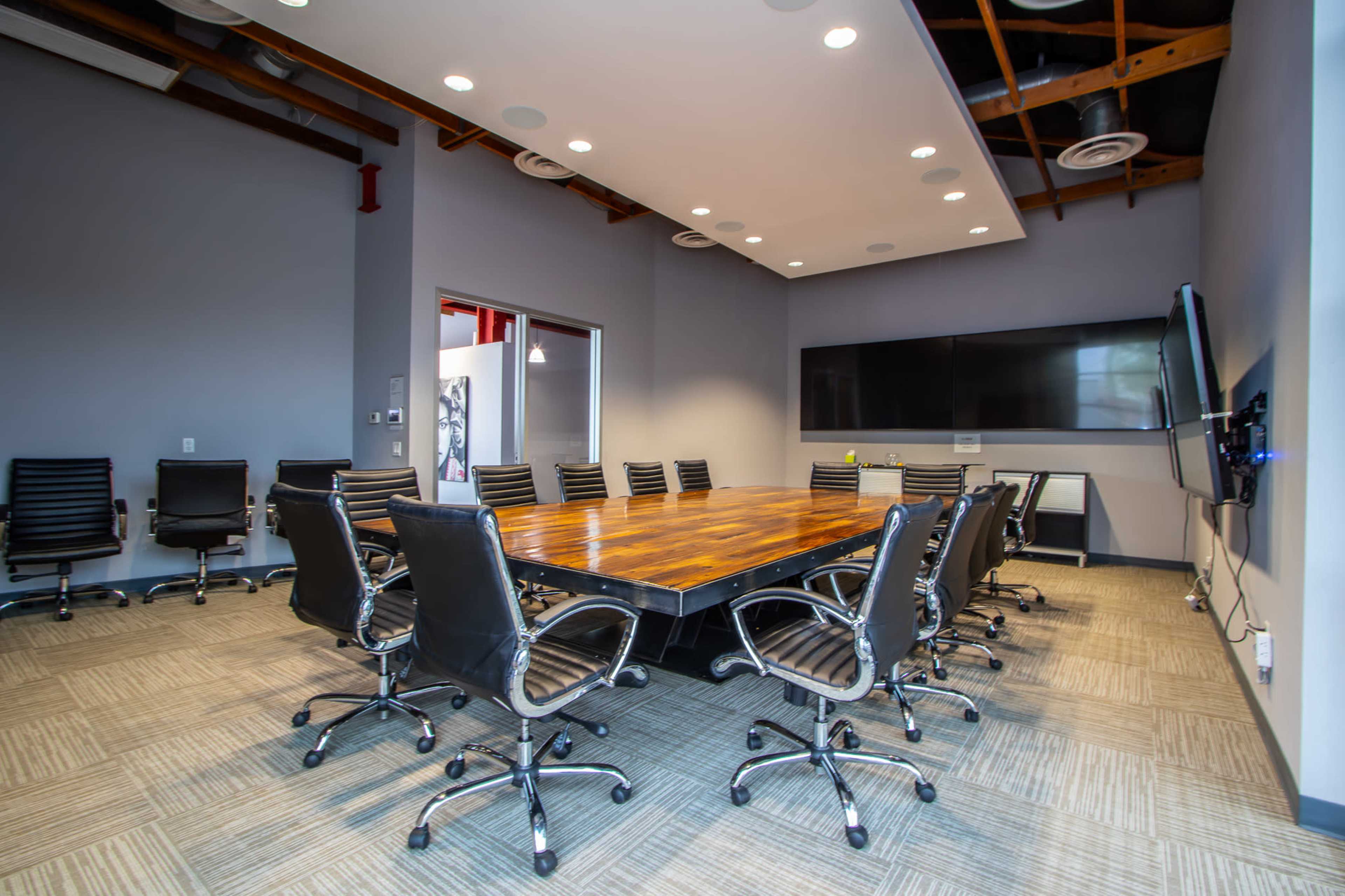 A large conference room features a rectangular wooden table surrounded by black rolling chairs, with a wall-mounted screen and modern lighting.