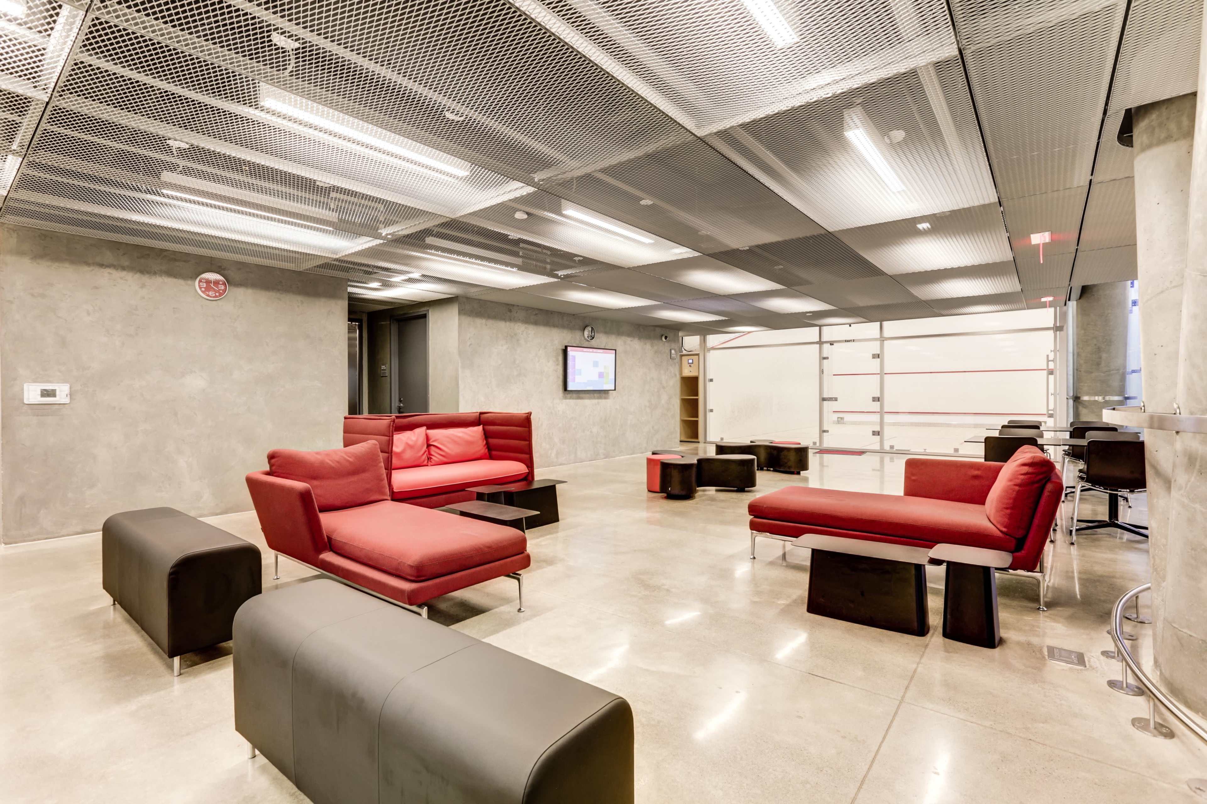 The image shows a modern lounge area with red sofas, black benches, and a sleek concrete floor, featuring large windows and overhead lighting.