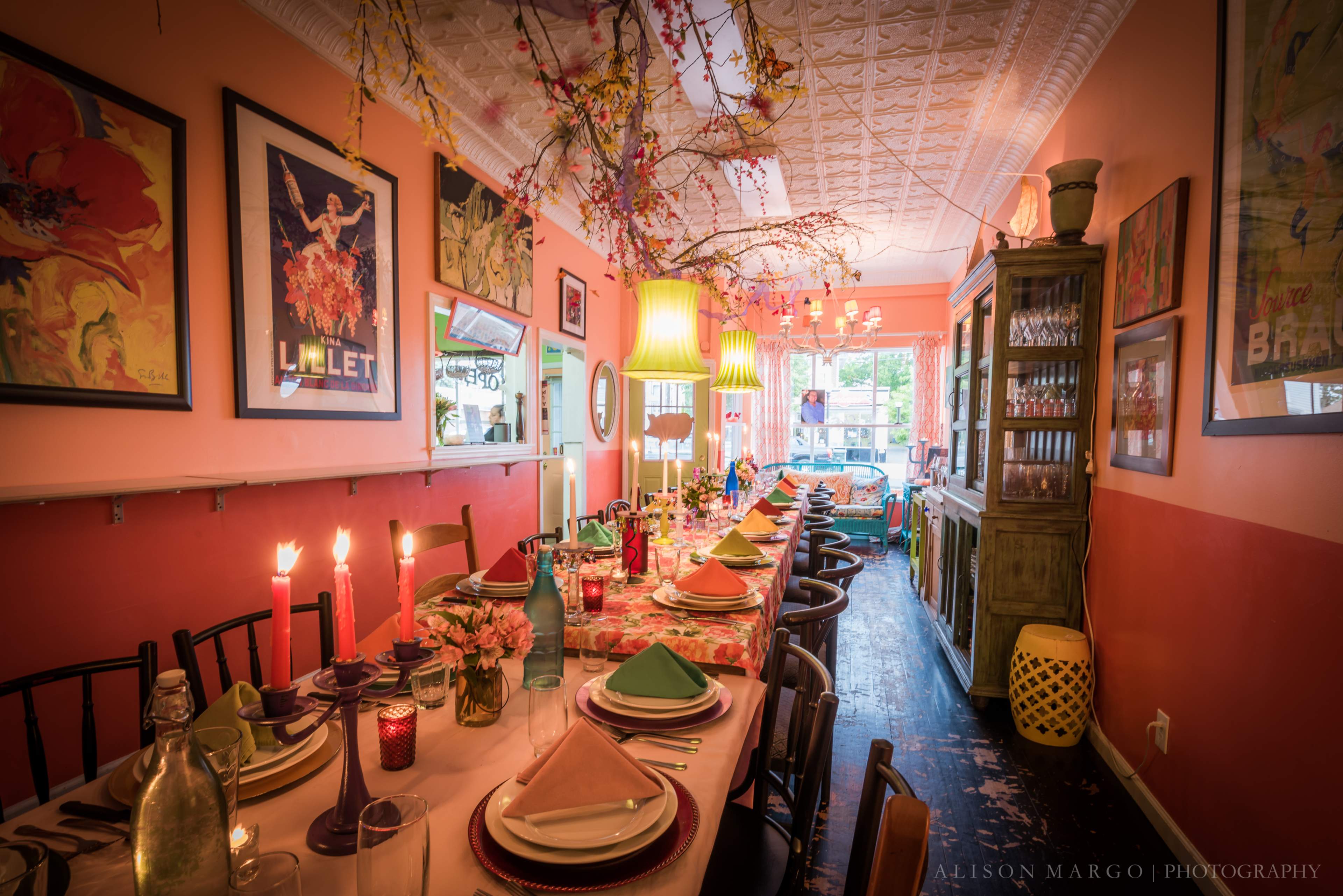 A long dining table is set with colorful tableware, surrounded by chairs, in a warmly decorated room featuring floral artwork and overhead decorations.