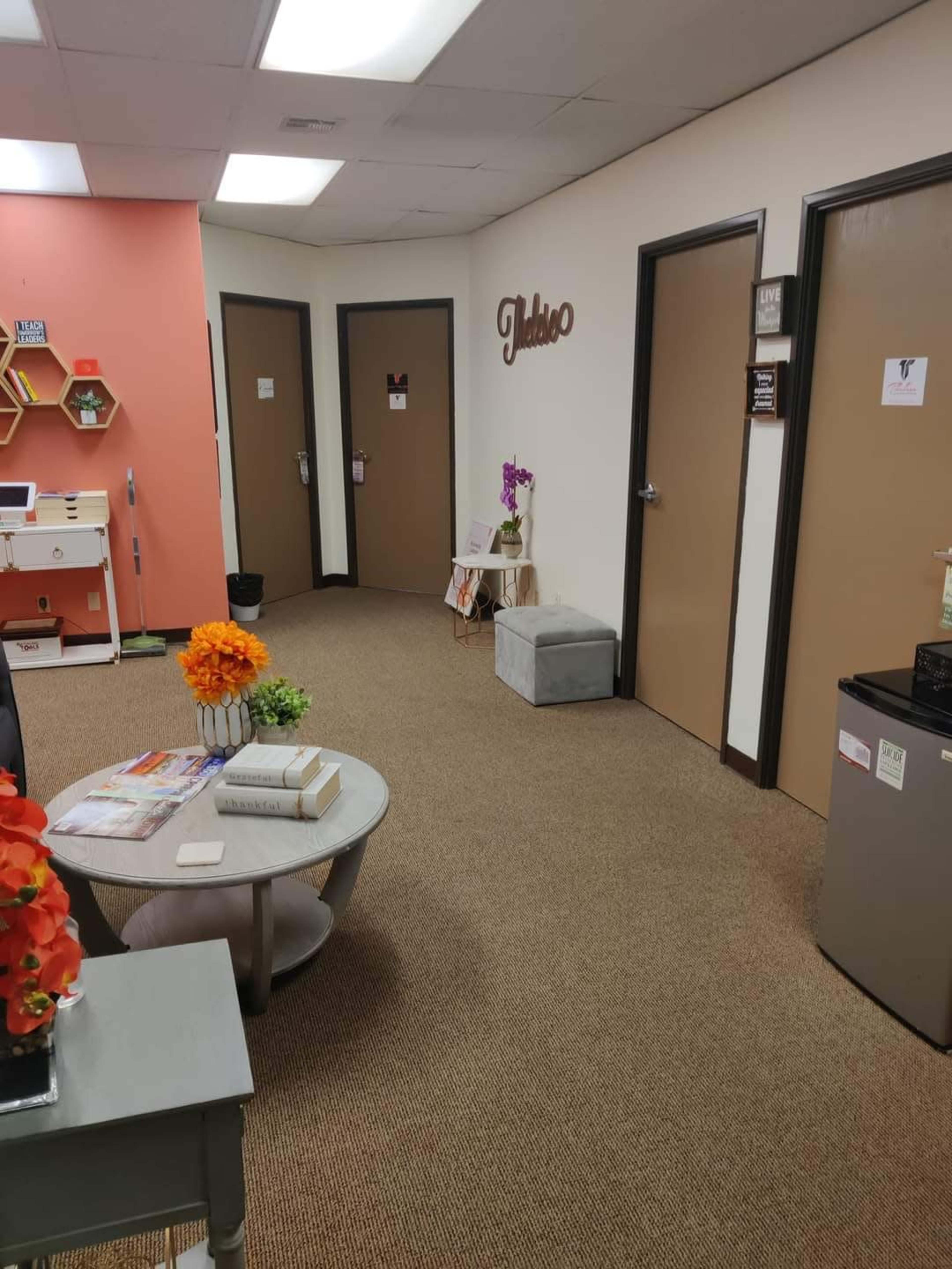A hallway with beige carpet, featuring three closed doors, a small decorative table with a flower arrangement, and a seating area with a round table and magazines.