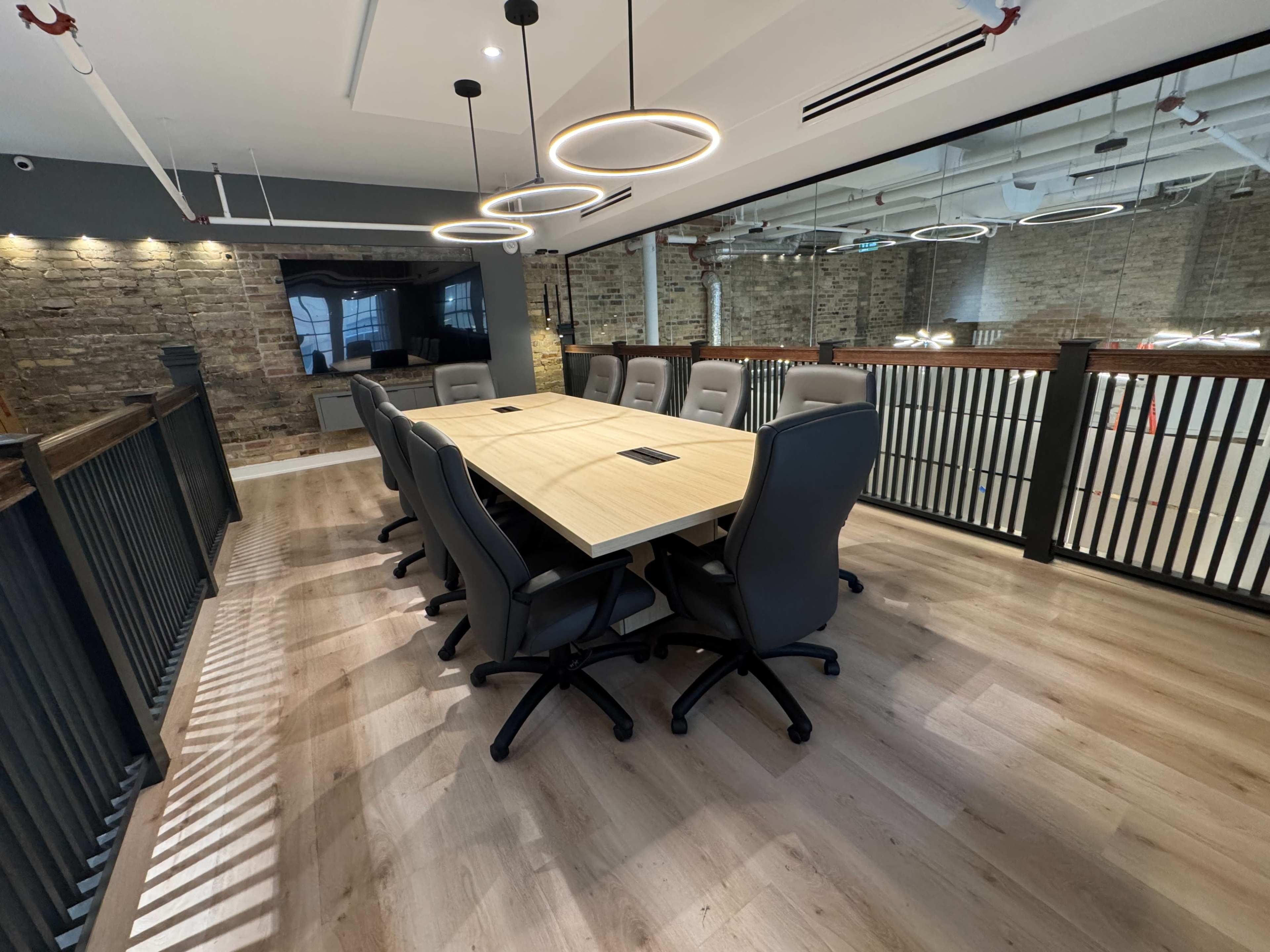 The image shows a modern conference room featuring a large rectangular table surrounded by gray rolling chairs, with a rustic stone wall and overhead lighting.