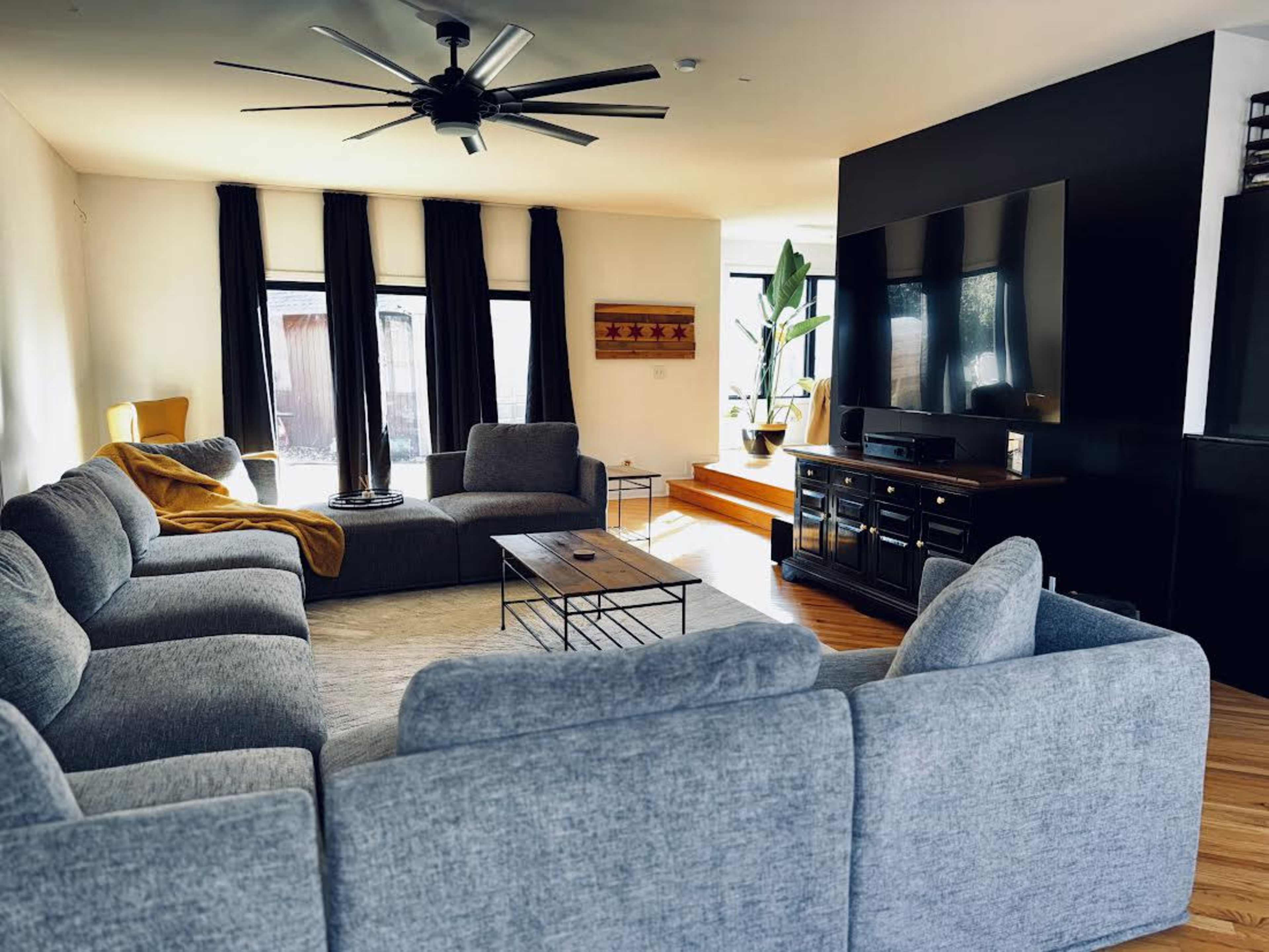 A spacious living room features a large gray sectional sofa, a wooden coffee table, and a wall-mounted television, with large windows allowing natural light to enter the space.