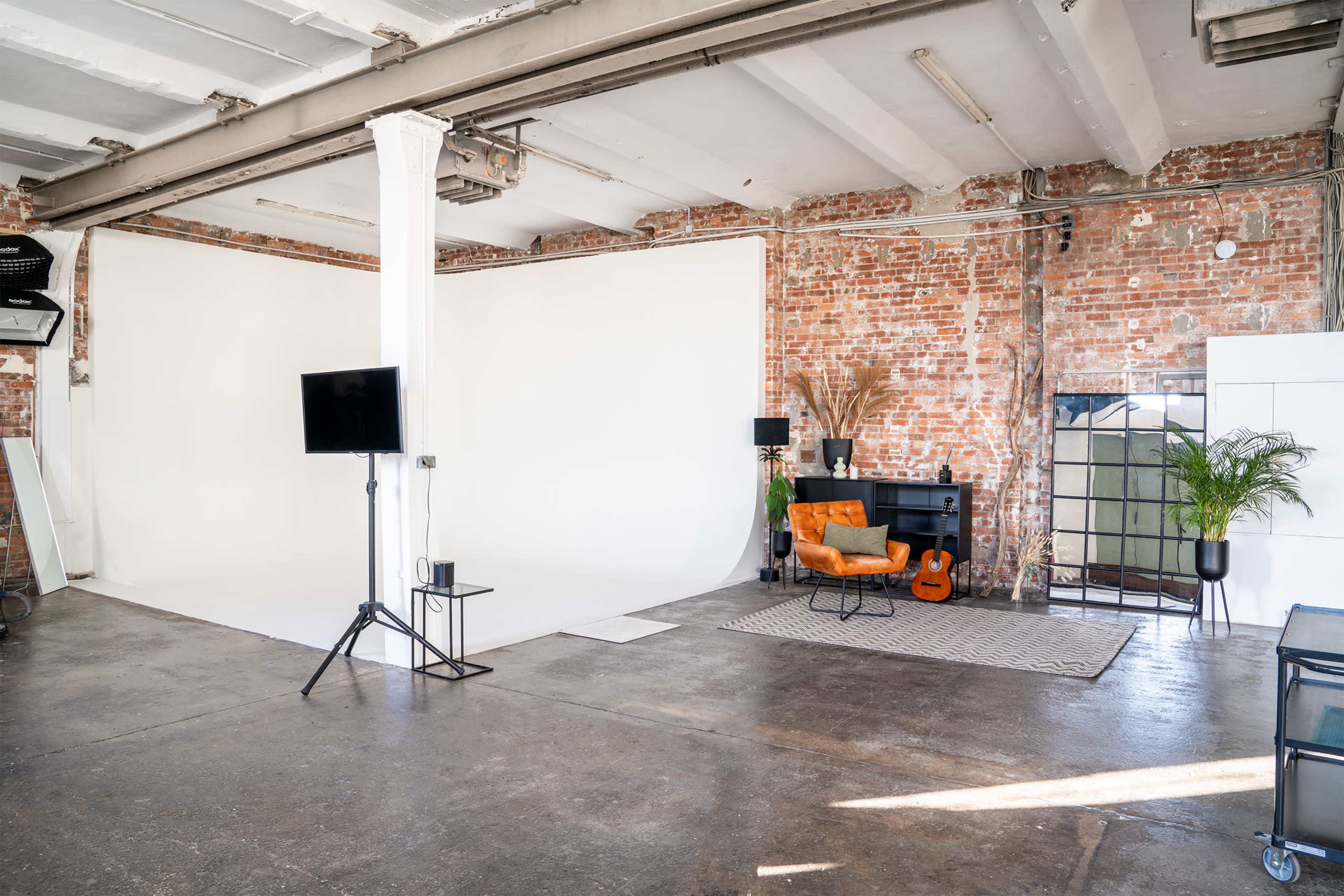 The image shows a spacious studio with a white backdrop, exposed brick walls, and a cozy seating area featuring an orange chair, a guitar, and potted plants.