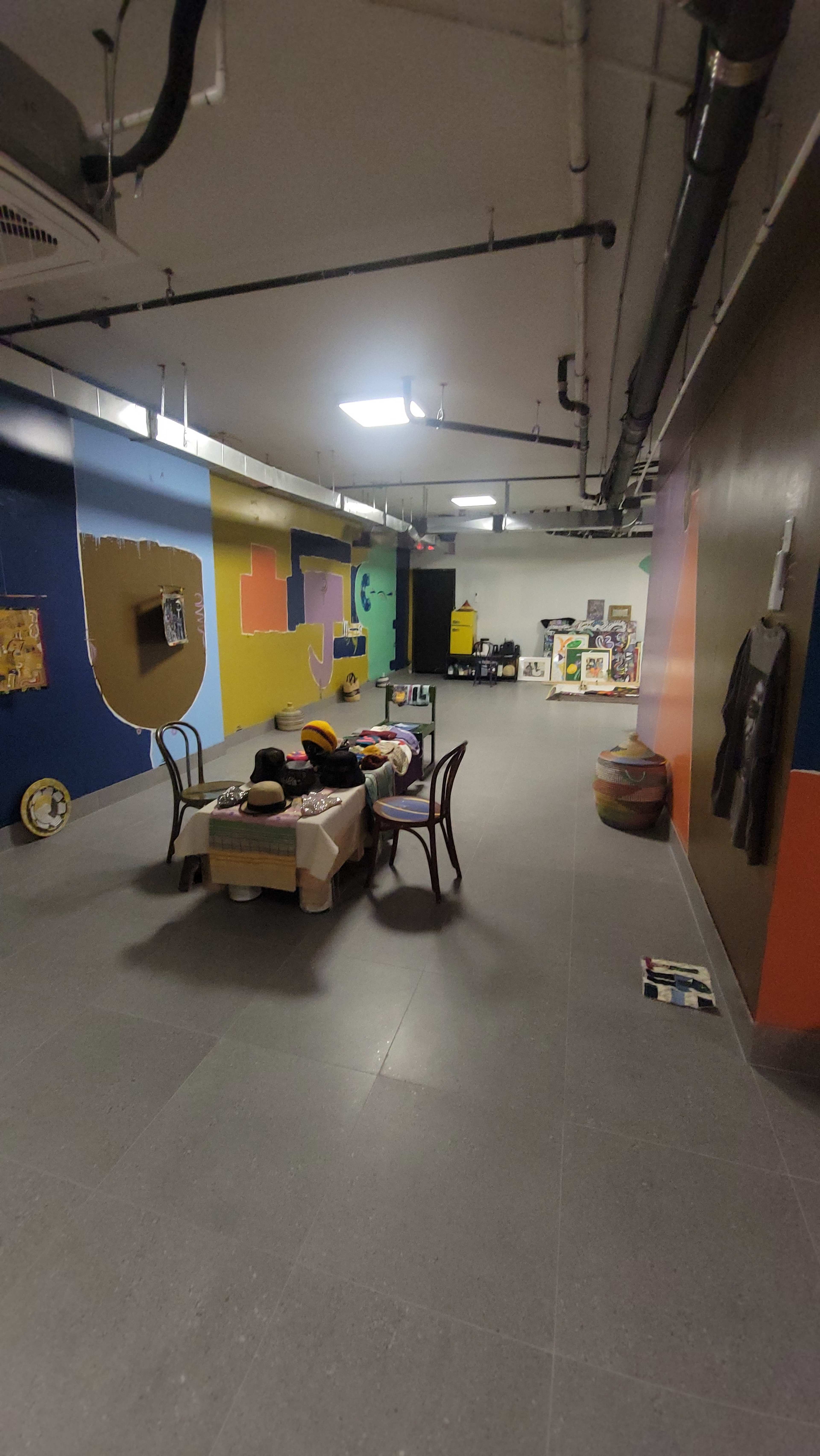The image shows a spacious basement room with colorful walls, a table in the center, and various objects scattered around, including chairs and artworks.