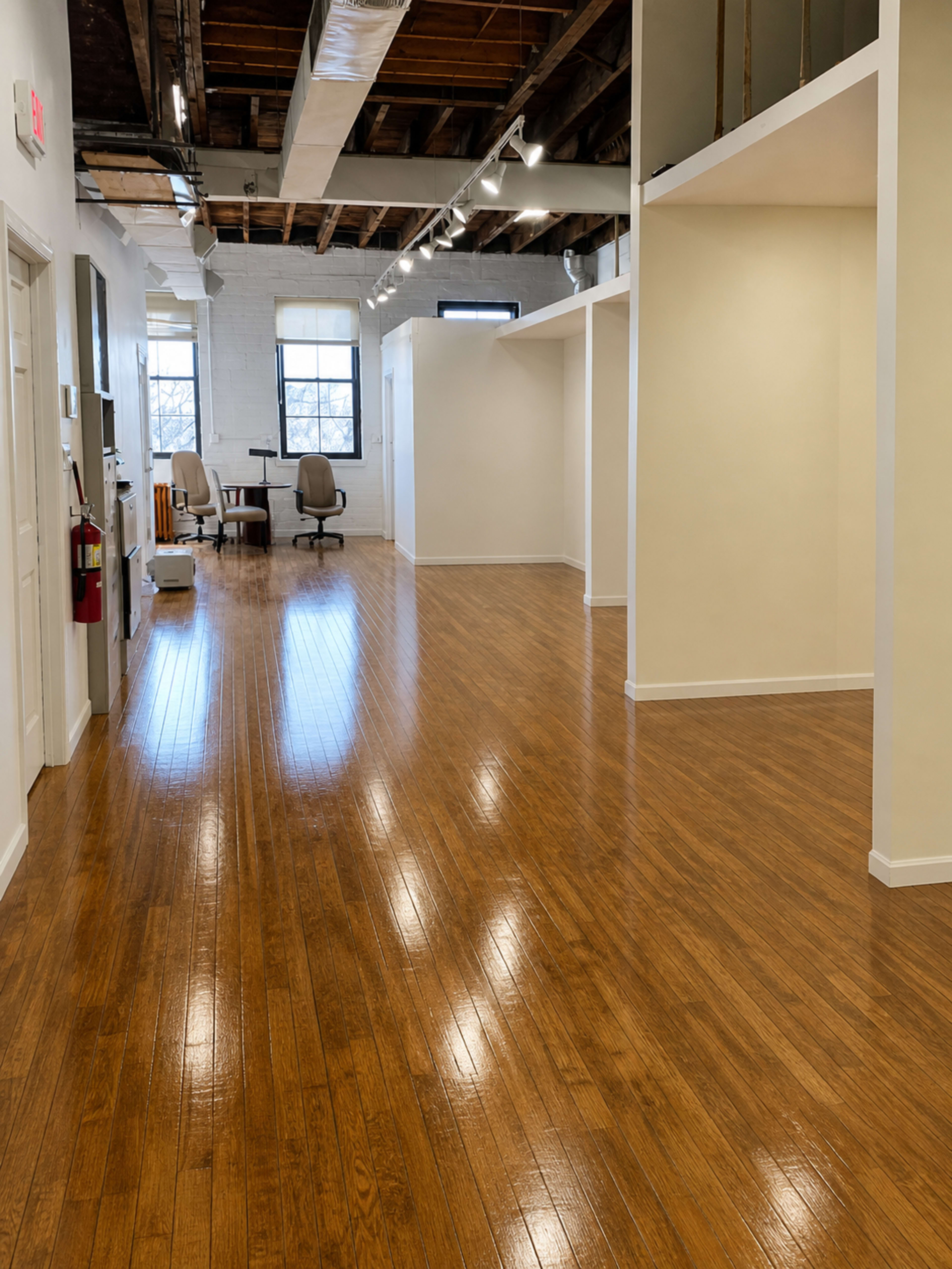 The image shows a spacious, well-lit interior of an office with polished wooden floors, white walls, and visible office furniture in the background.