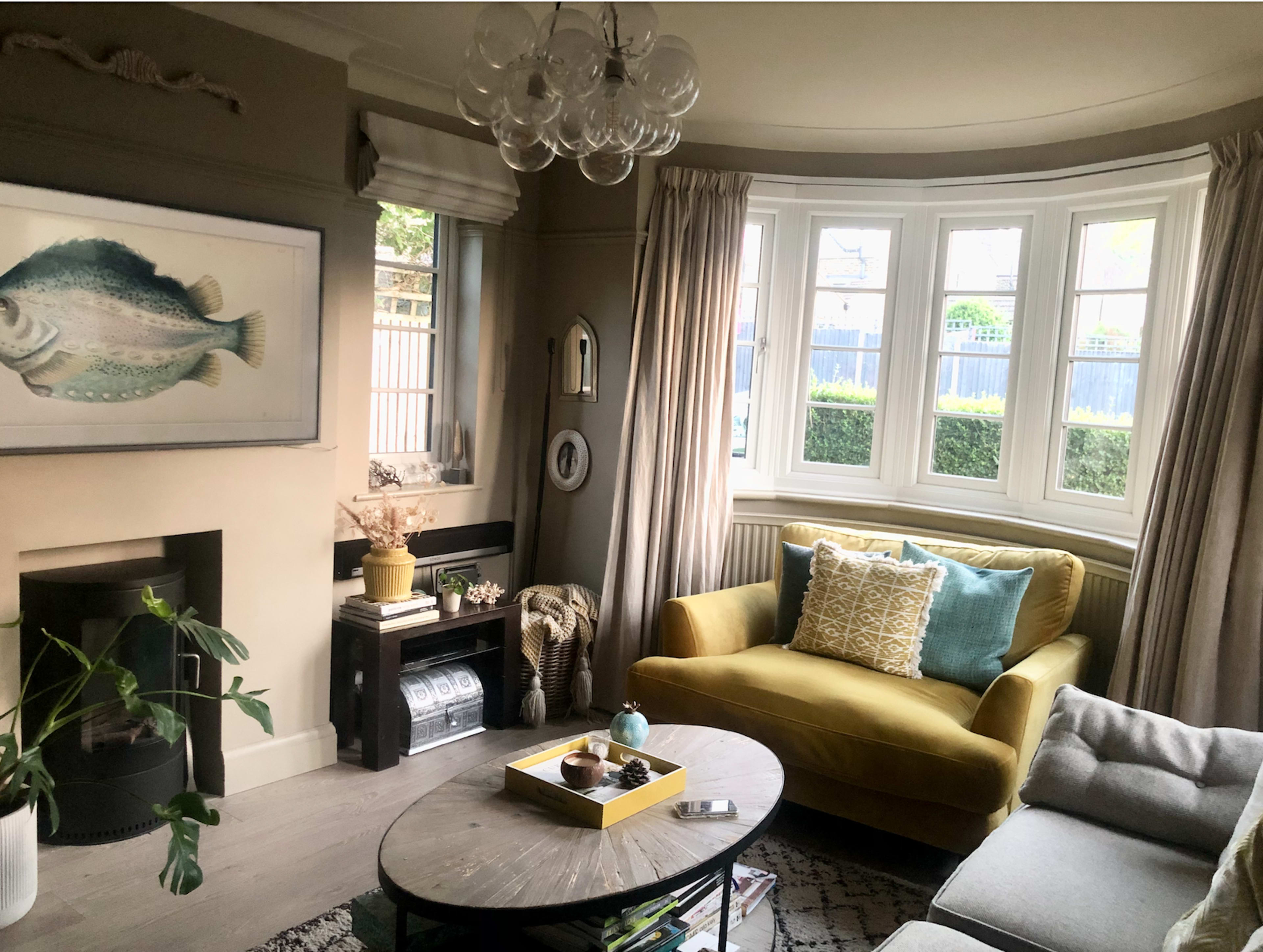 A cozy living room features a yellow sofa, a round coffee table, large windows, and decorative elements including a fish painting and potted plants.