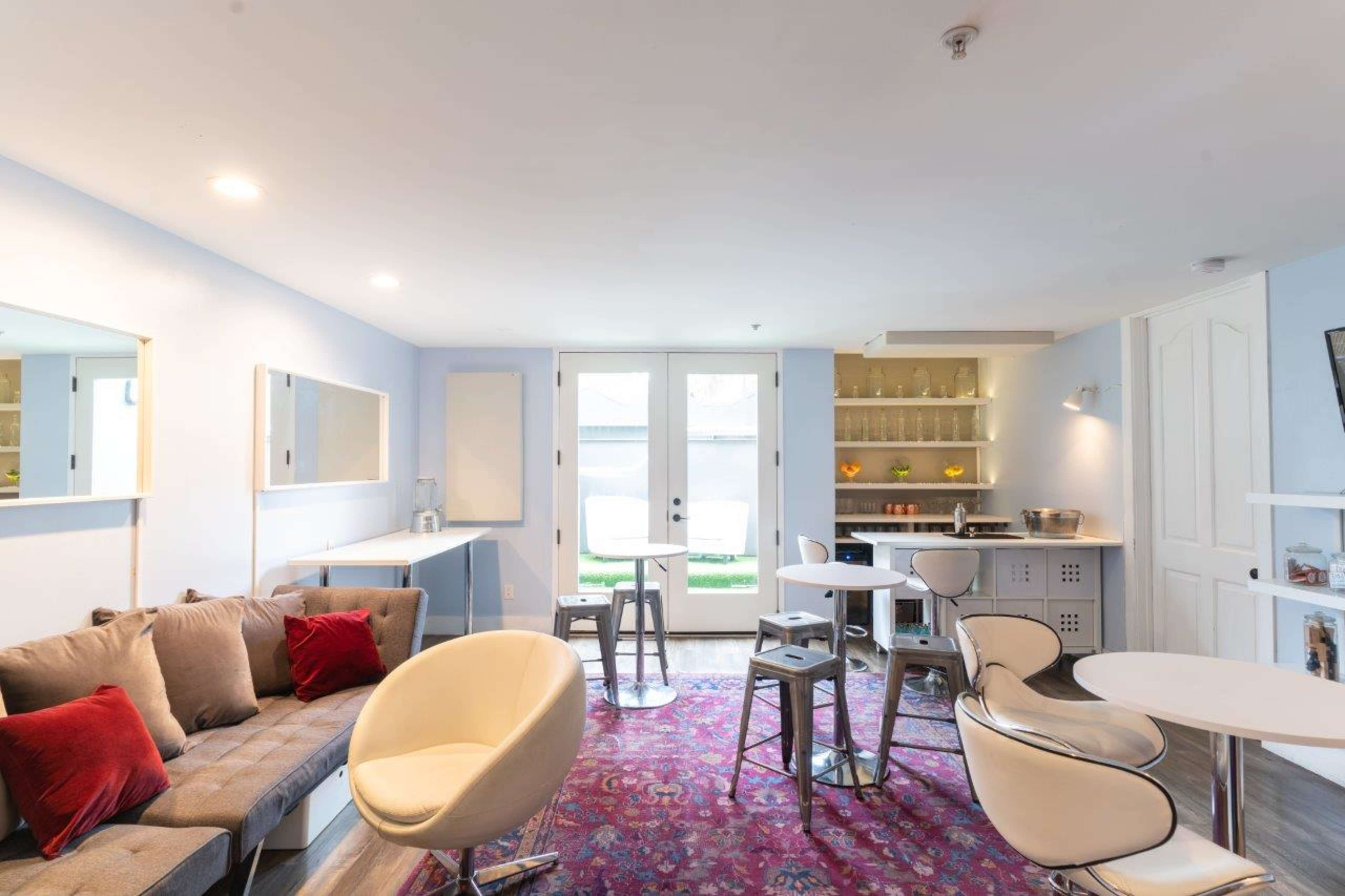 A modern lounge area features a gray sofa, white chairs, small tables, and a bar with glass shelves, leading to a doorway with access to an outdoor space.