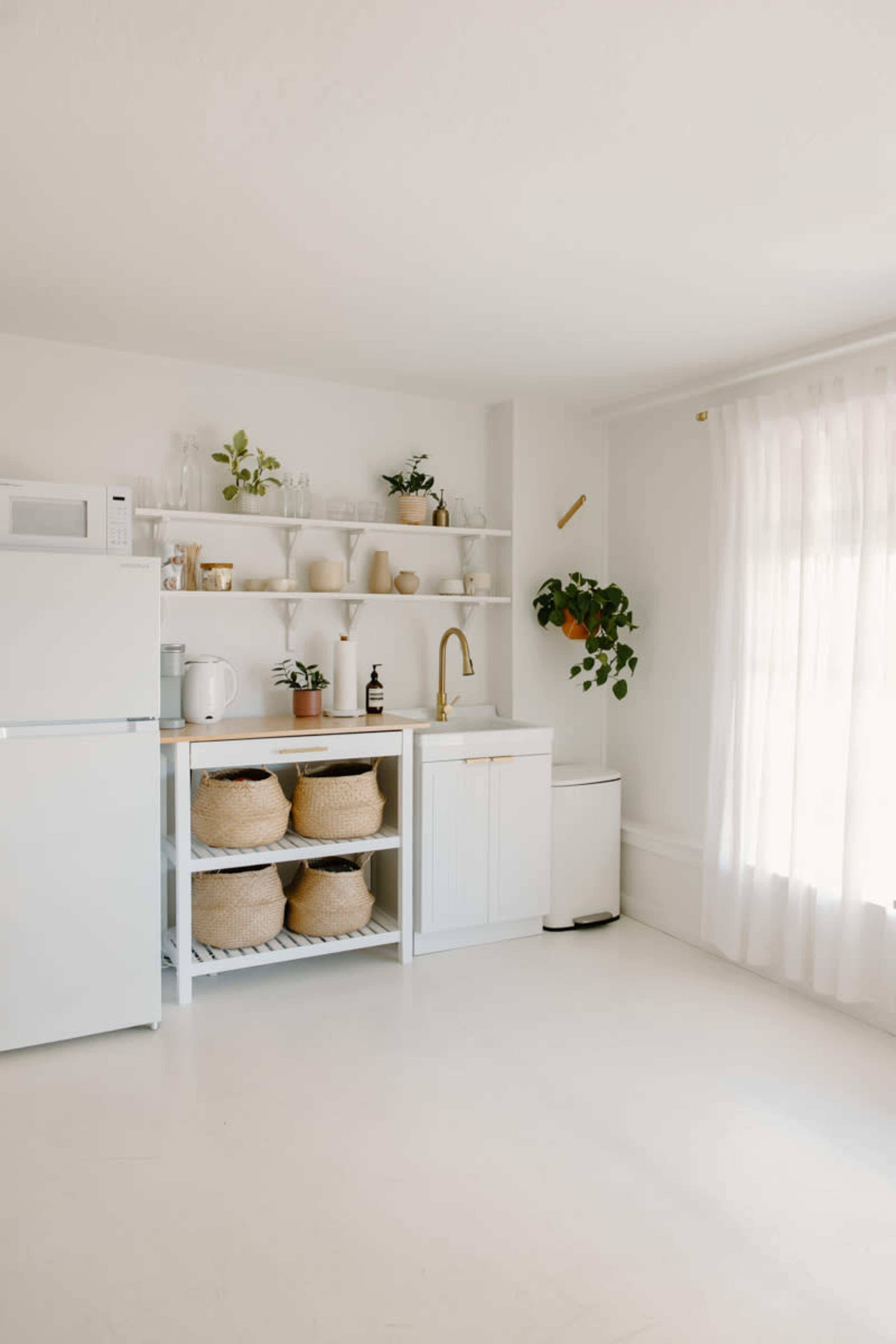 A modern kitchen features white cabinetry, open shelving with decorative items, a gold faucet, and potted plants, illuminated by natural light from a window.