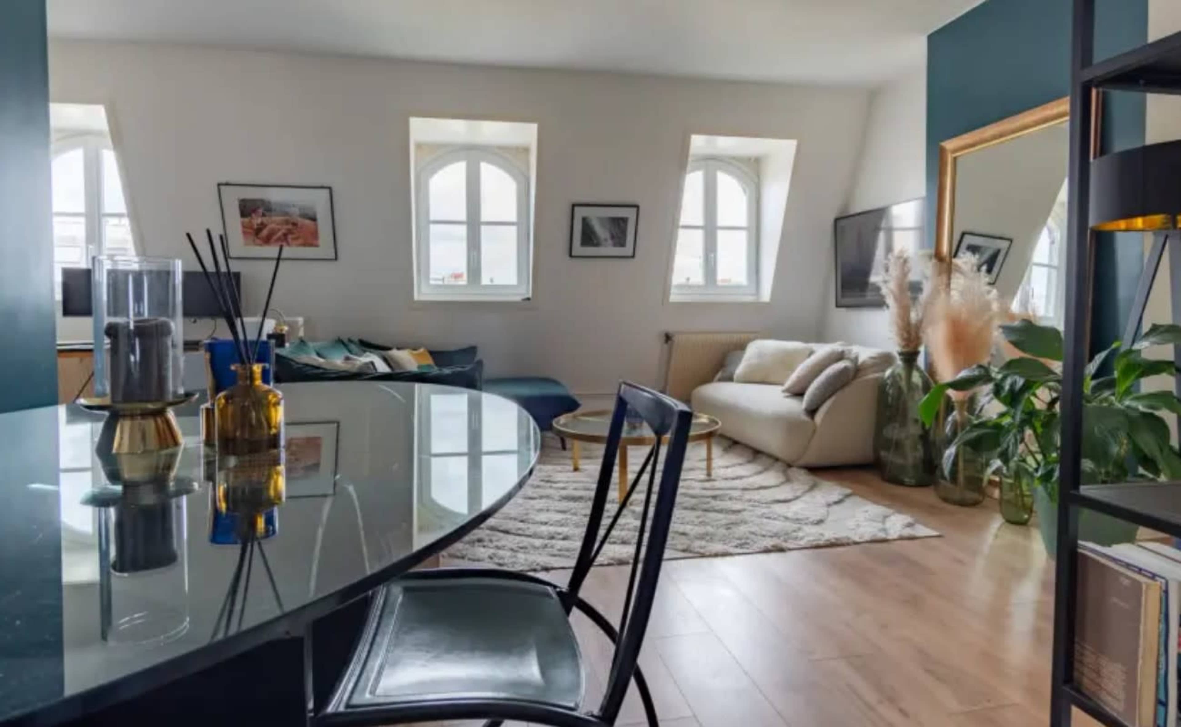 A bright, modern living room features a glass-top dining table, a light-colored sofa, and multiple windows that frame the view of the outdoors.