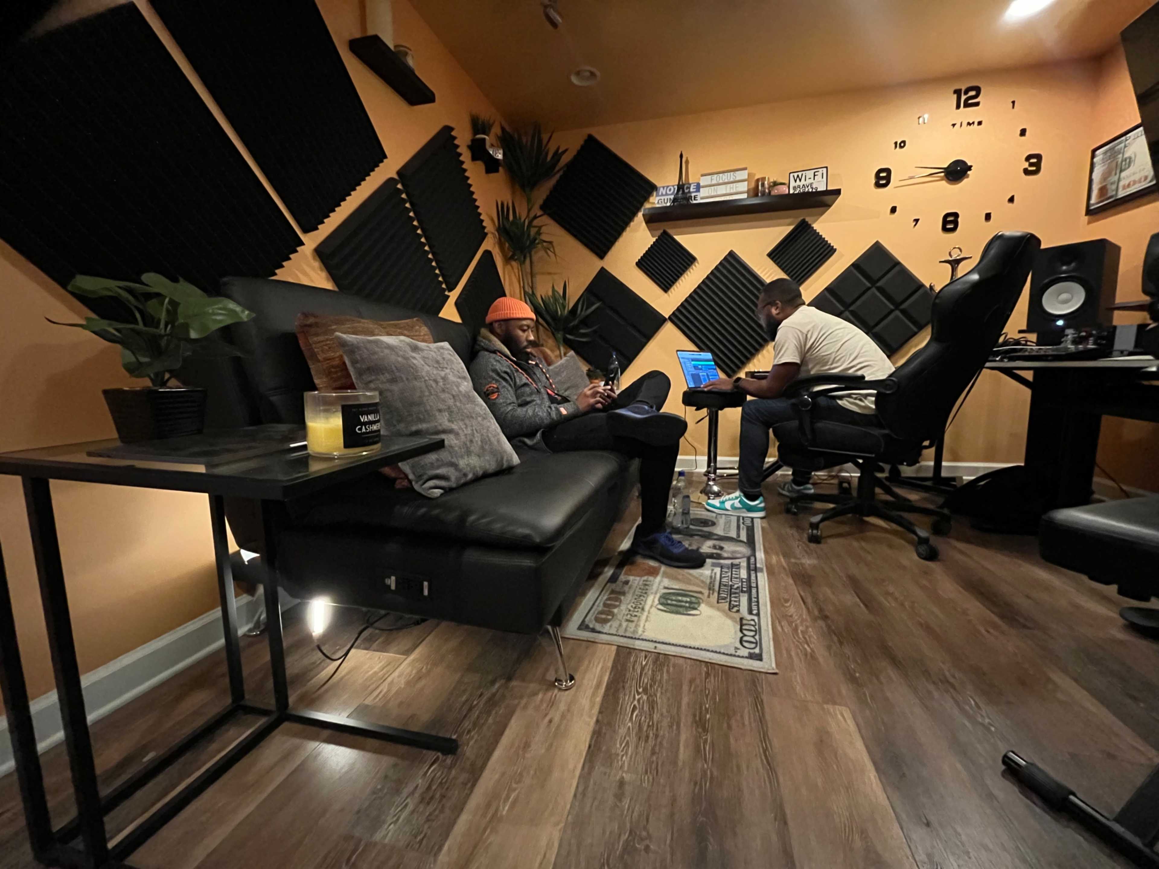 A cozy recording studio features a black leather couch, a person using a phone, and another individual working at a desk with recording equipment.