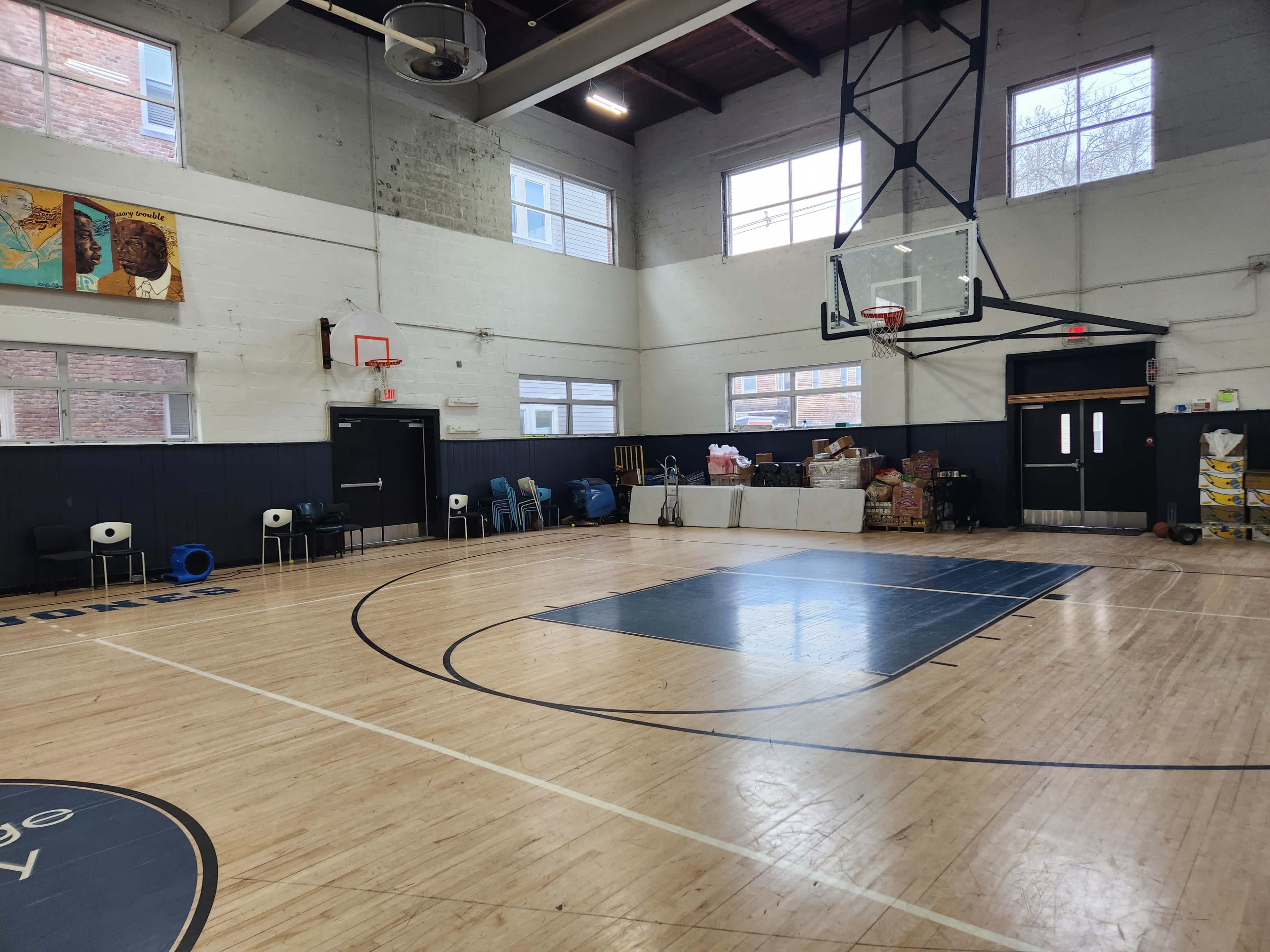 The image shows an indoor basketball court with a hoop and some seating along one side, along with boxes and supplies stacked in the background.