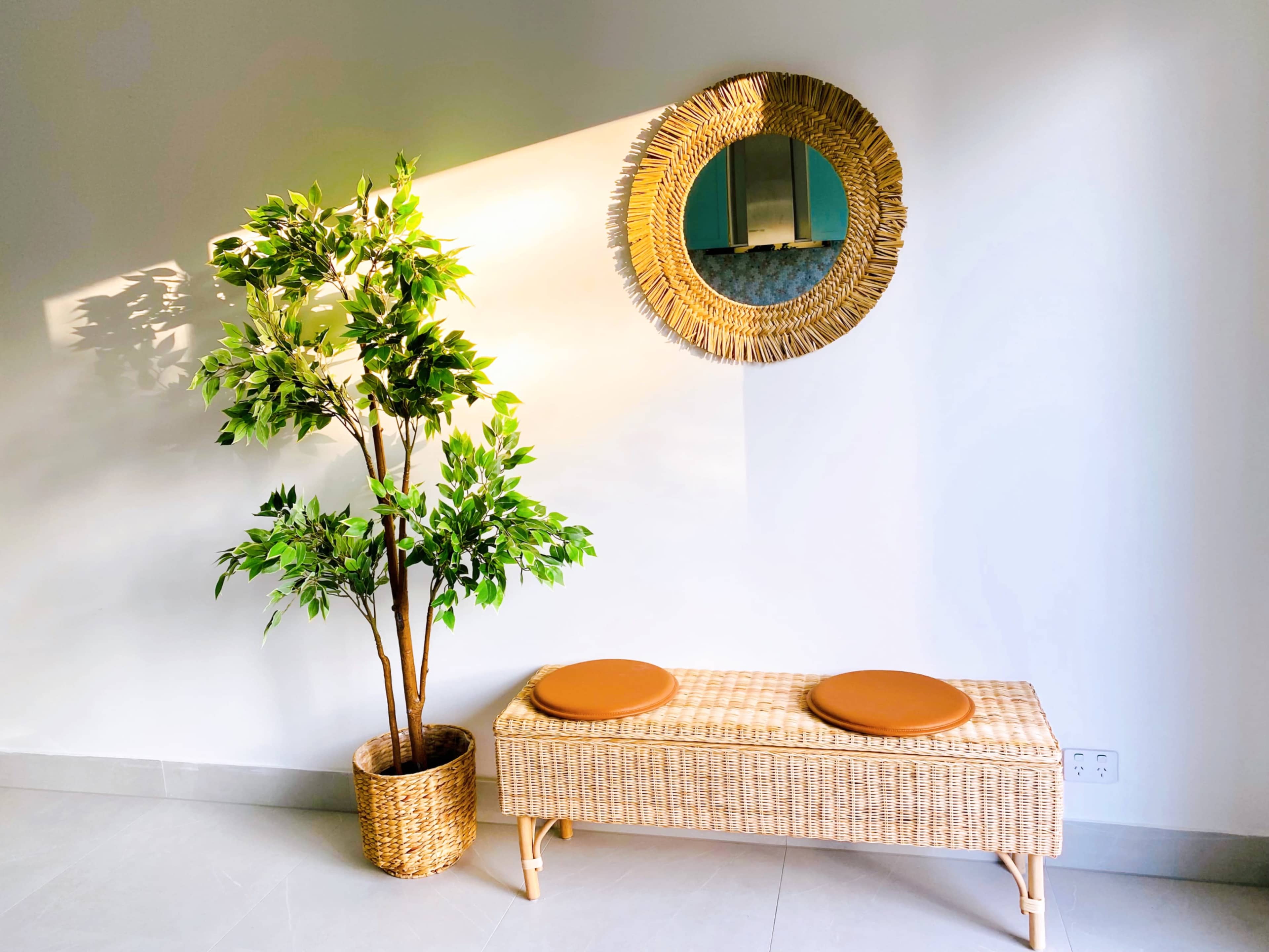 A wicker bench with two round cushions sits beside a potted plant and a circular mirror on a light-colored wall.