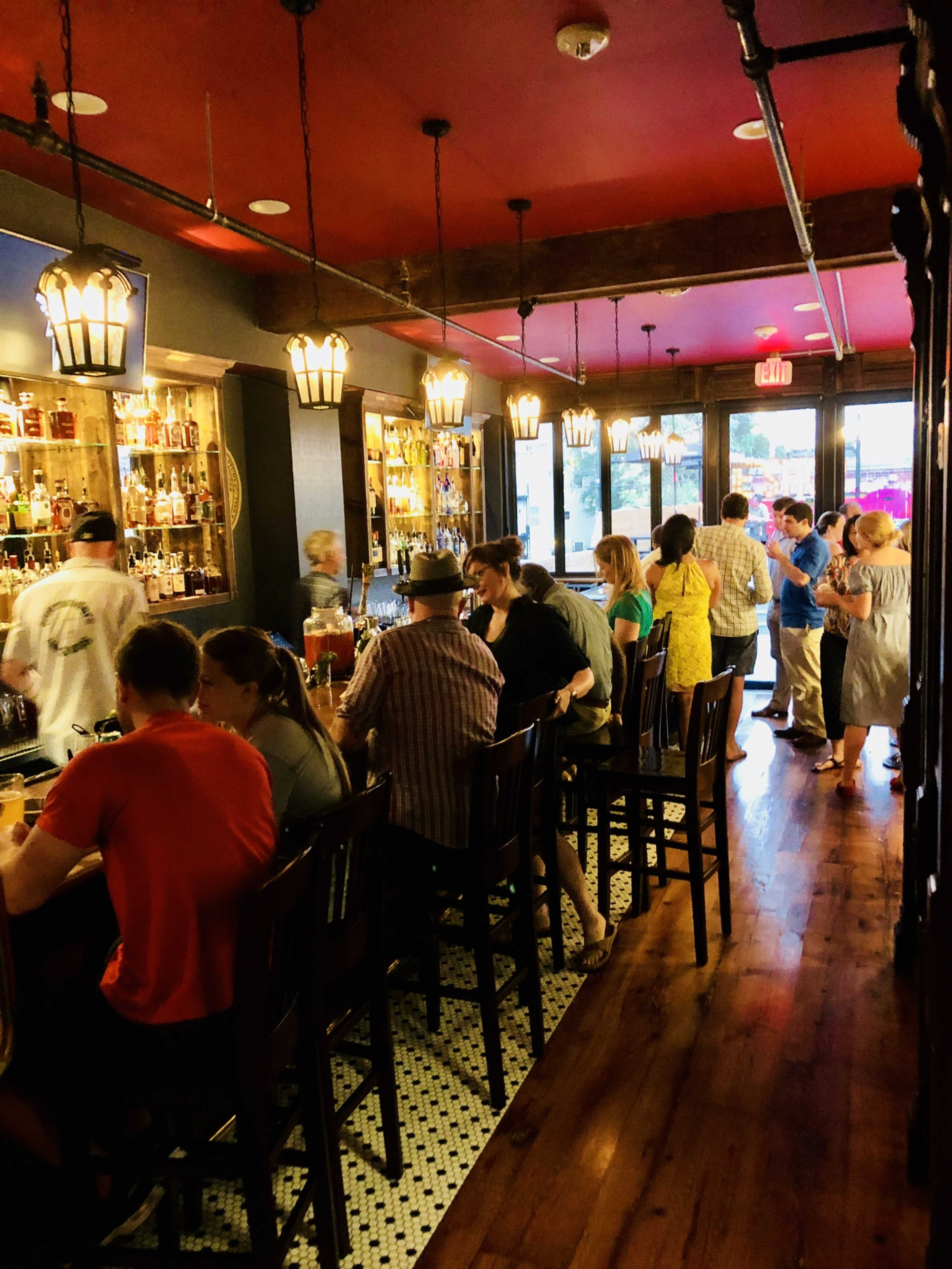 The image shows a busy bar interior with patrons seated at a long counter and others standing near shelves filled with bottles.