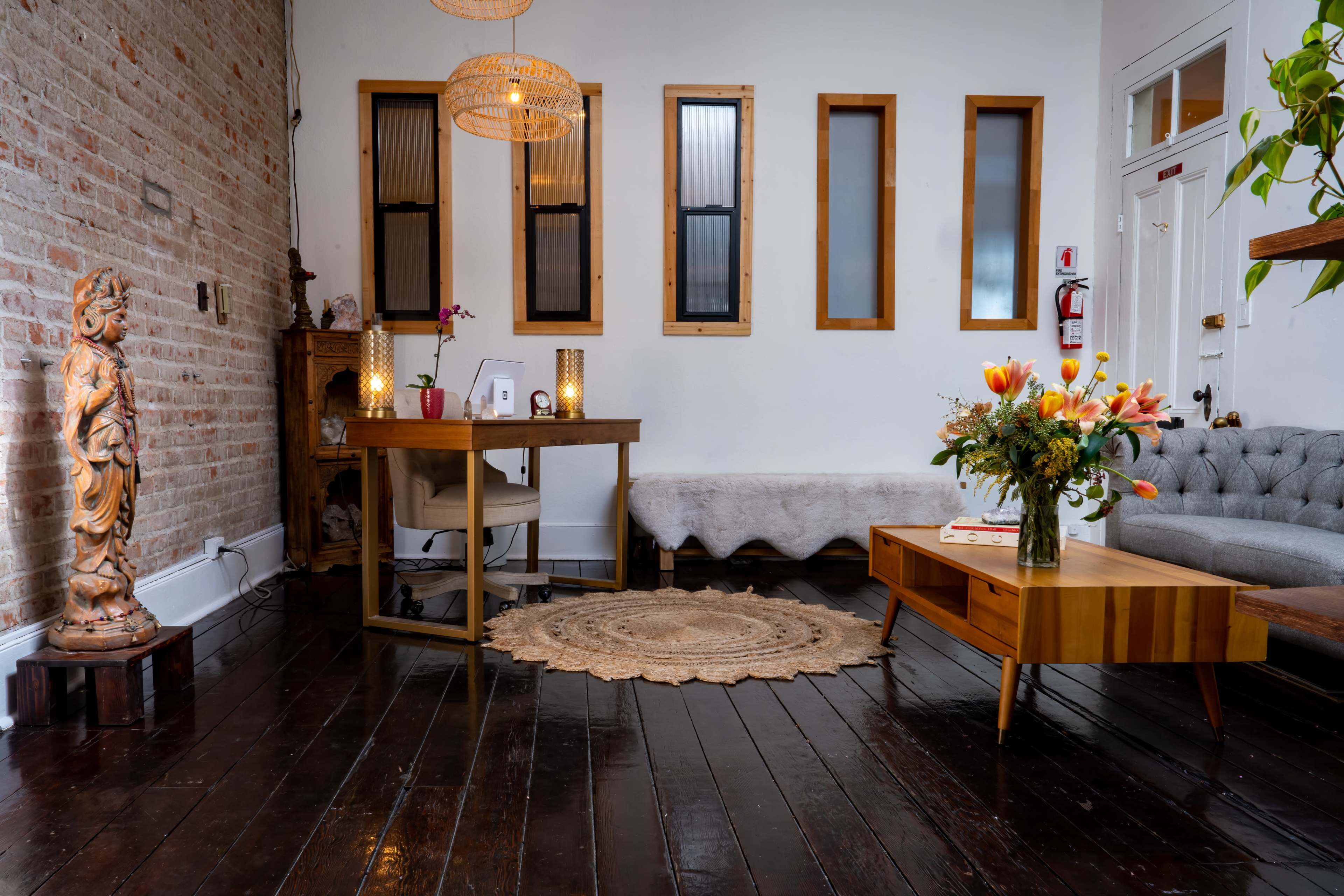 A cozy interior space with a wooden desk, a gray couch, decorative plants, and a statue, all set against a brick wall.