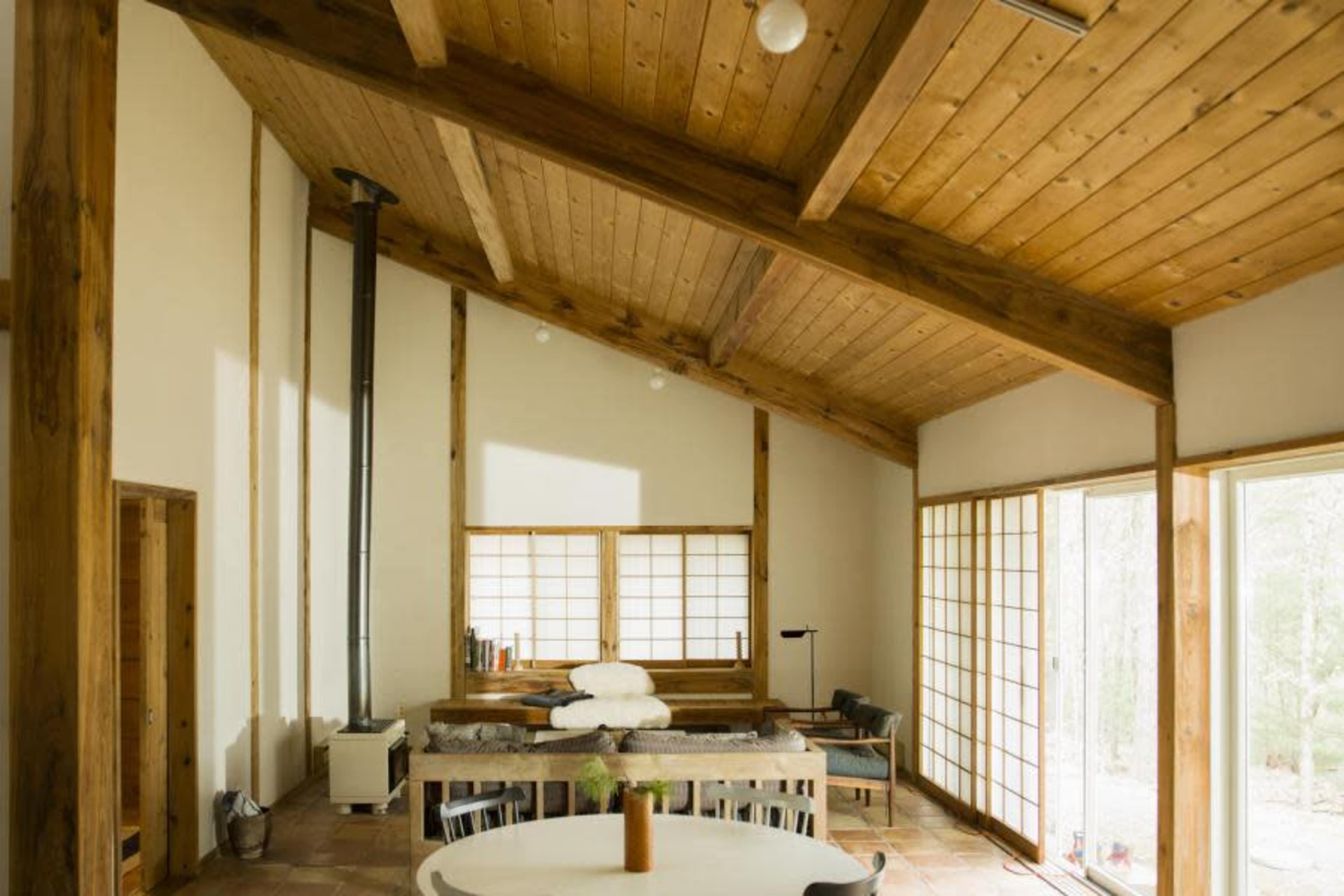 The image shows a modern interior of a house with wooden beams, a central dining table, and large windows that open to a natural setting.