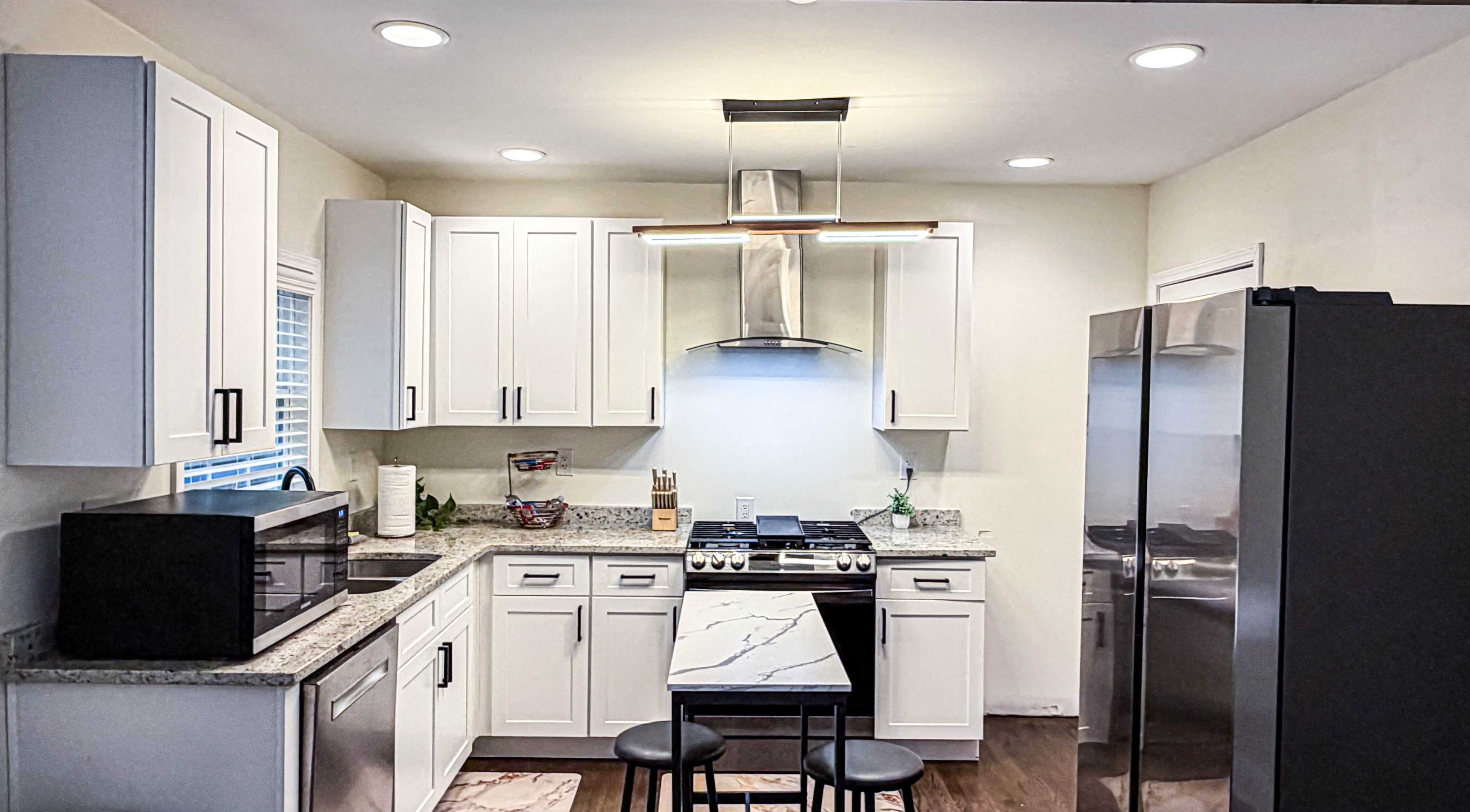 The image shows a modern kitchen with white cabinets, granite countertops, stainless steel appliances, and a central island with two black stools.