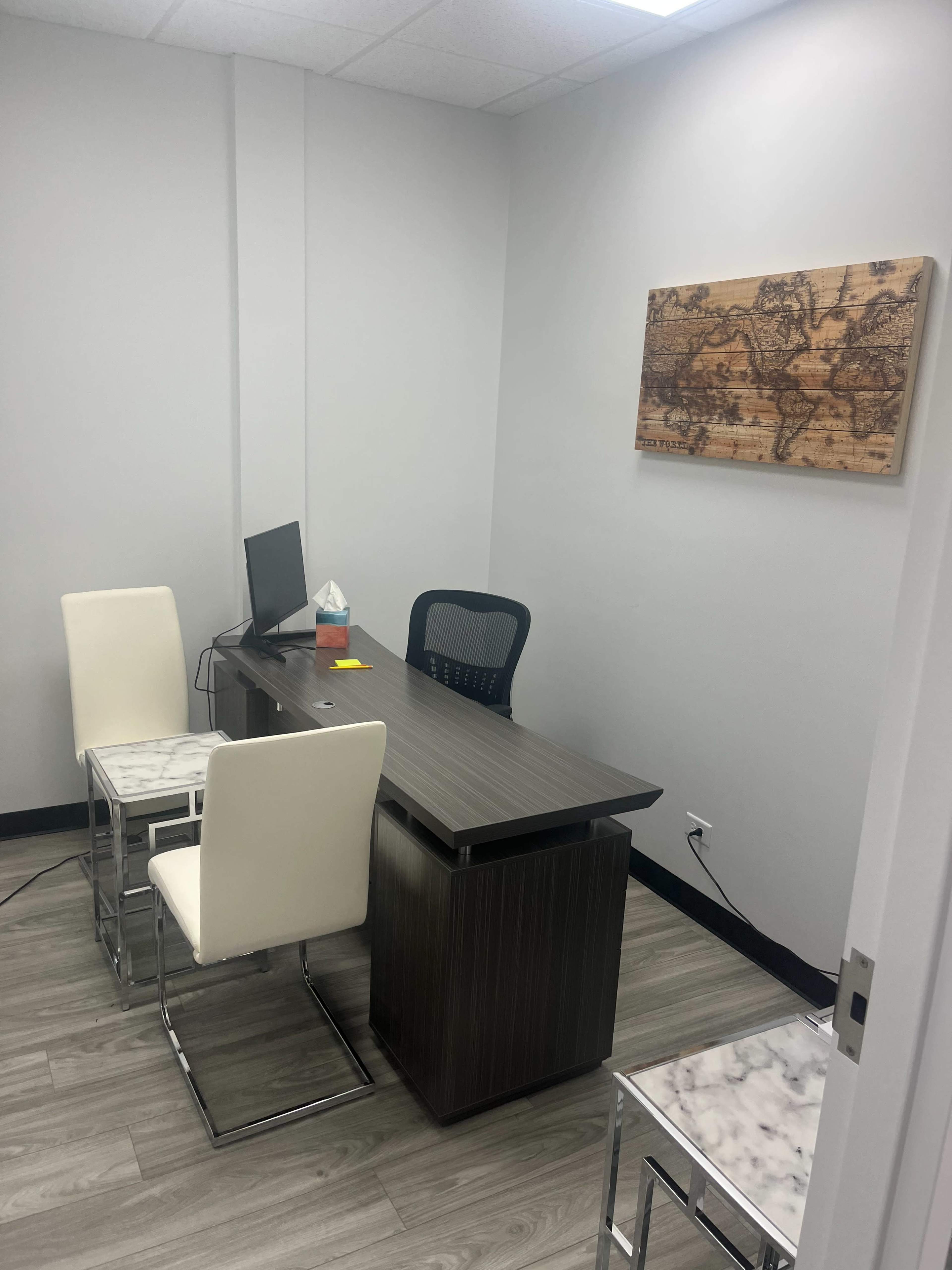 The image shows a small office space with a desk, two white chairs, a computer monitor, and a wooden world map on the wall.