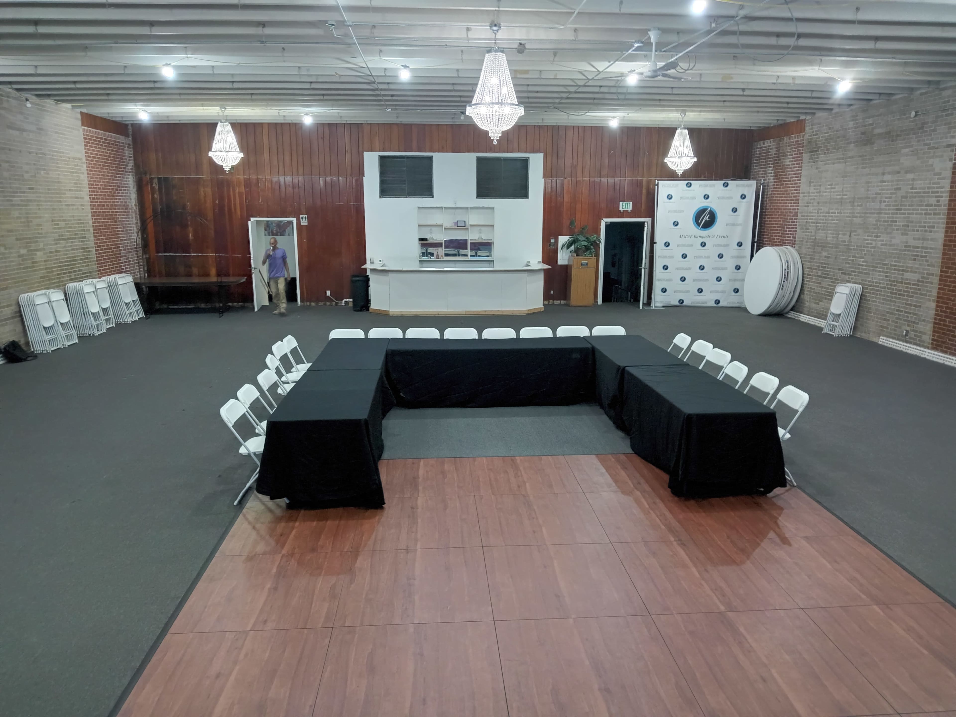A spacious indoor venue set up with a U-shaped arrangement of tables covered in black cloth, surrounded by white folding chairs, with a bar area and a backdrop featuring logos in the background.