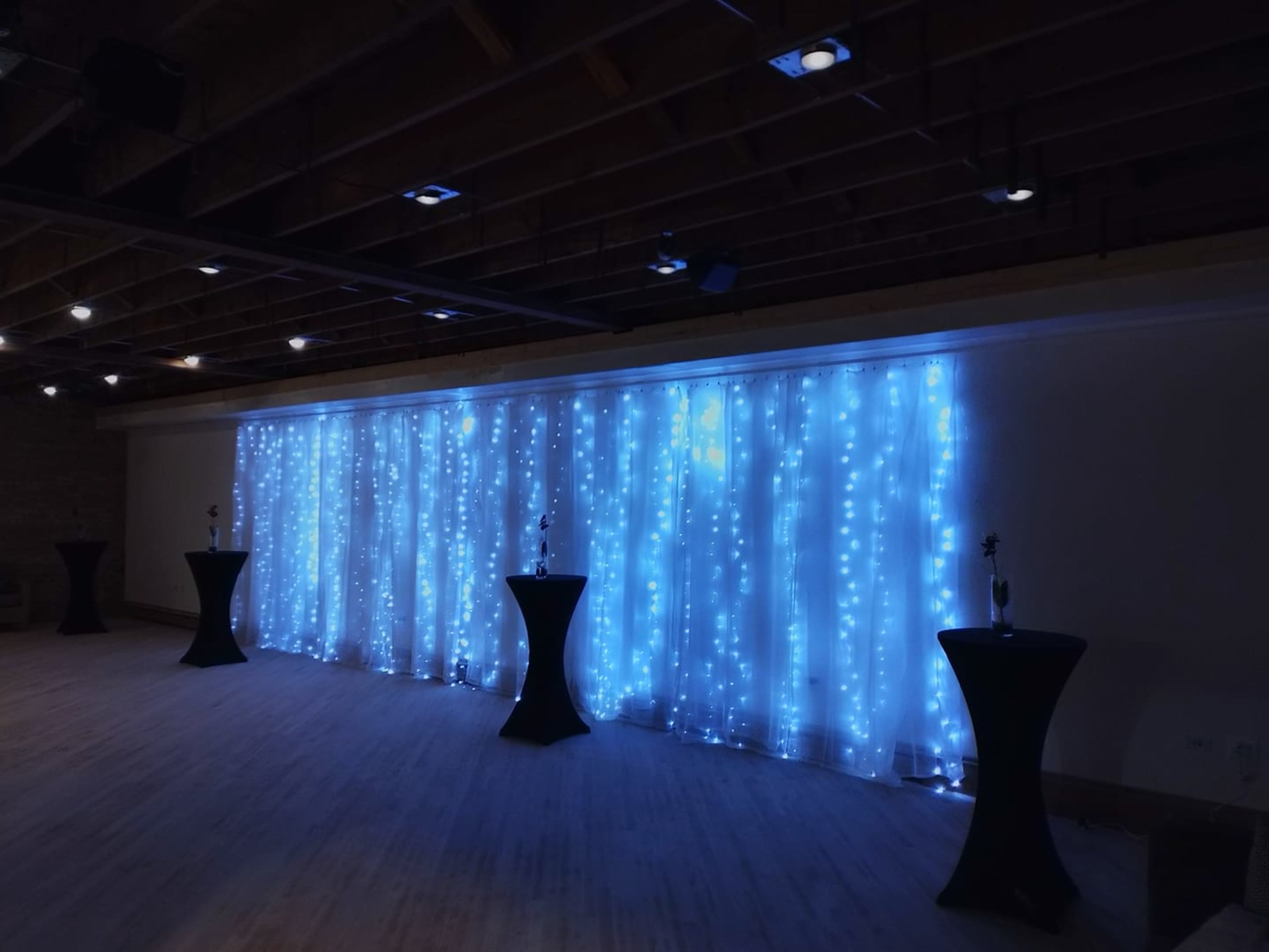 A room with a backdrop of blue fairy lights draped on a wall, accompanied by several black cocktail tables.