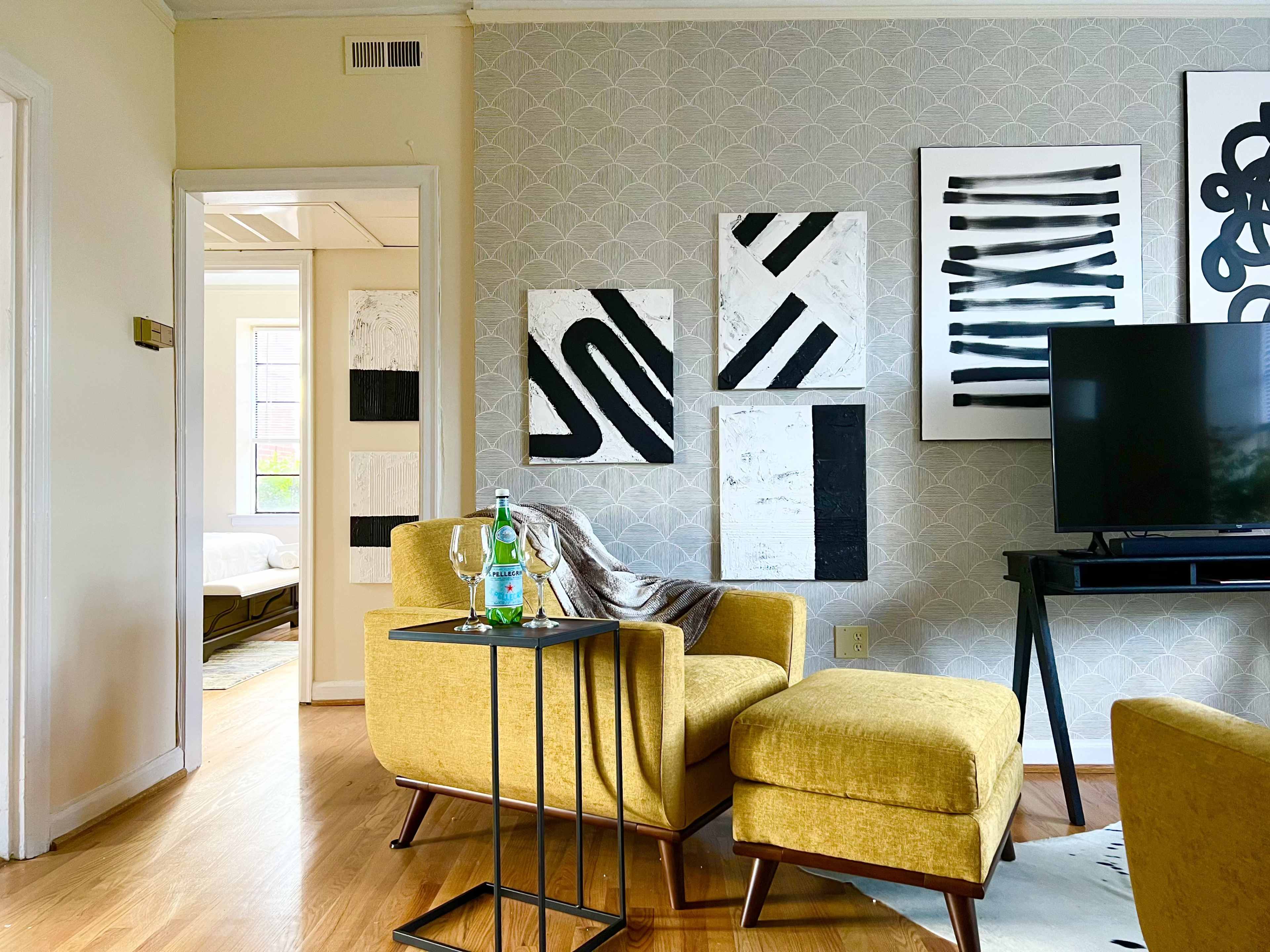 The scene features a modern living room with a yellow armchair and side table, black and white wall art, and a television, leading to a brightly lit room in the background.