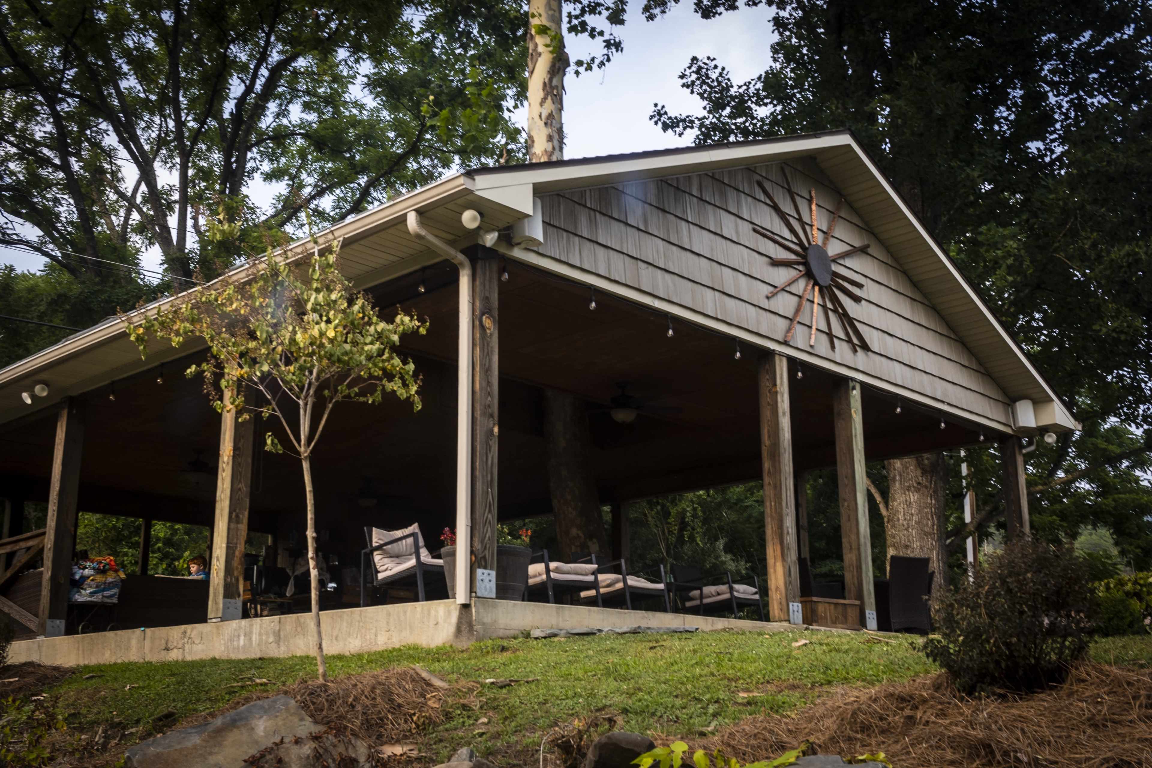 A covered pavilion with a large sunburst decoration is supported by wooden posts and surrounded by green foliage.