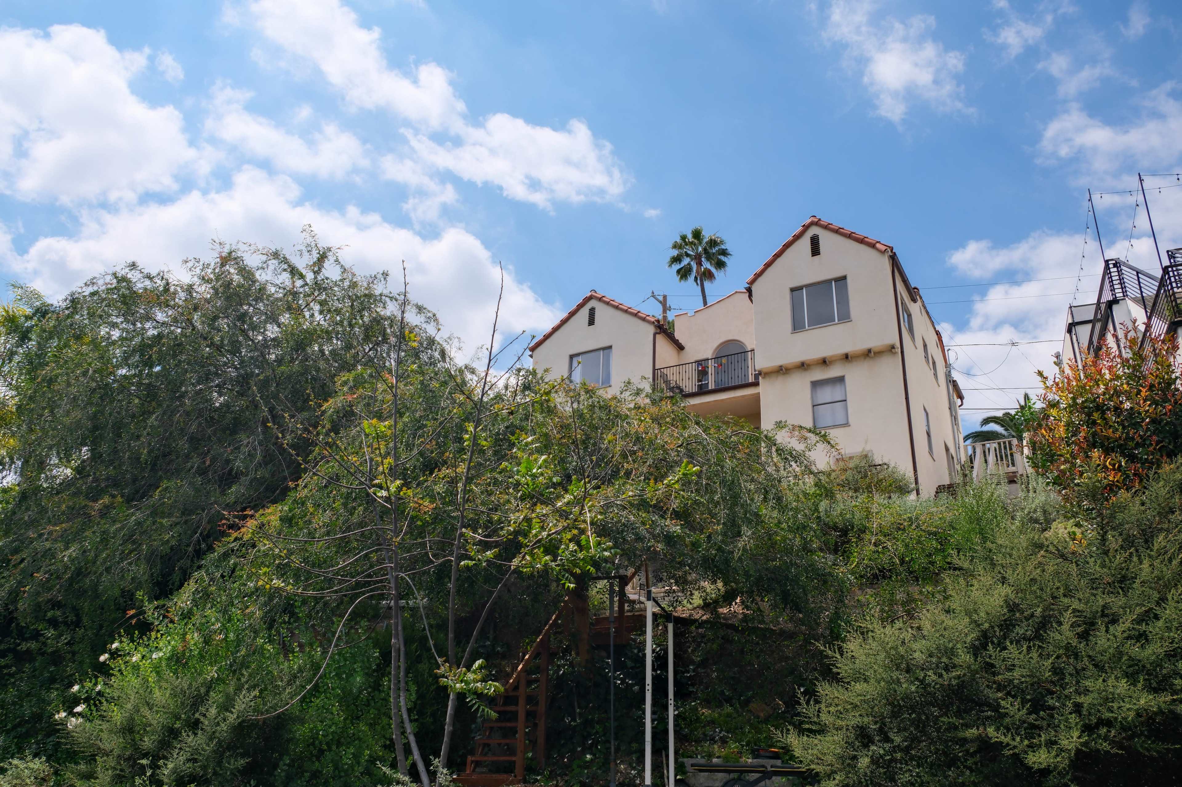 A two-story house with a sloped roof and a balcony is surrounded by lush greenery and a clear blue sky.