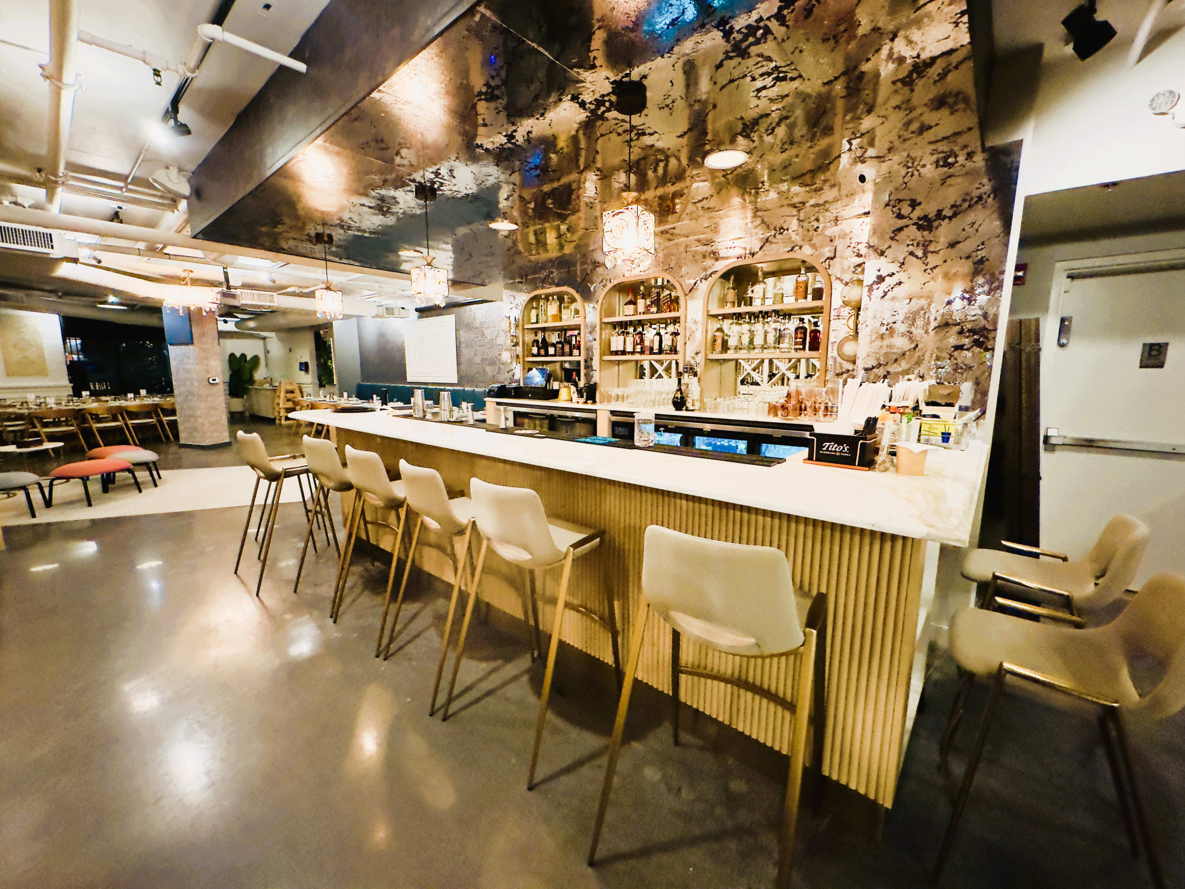 A modern bar area features a long counter with high stools, framed by decorative mirrors and shelves filled with bottles.