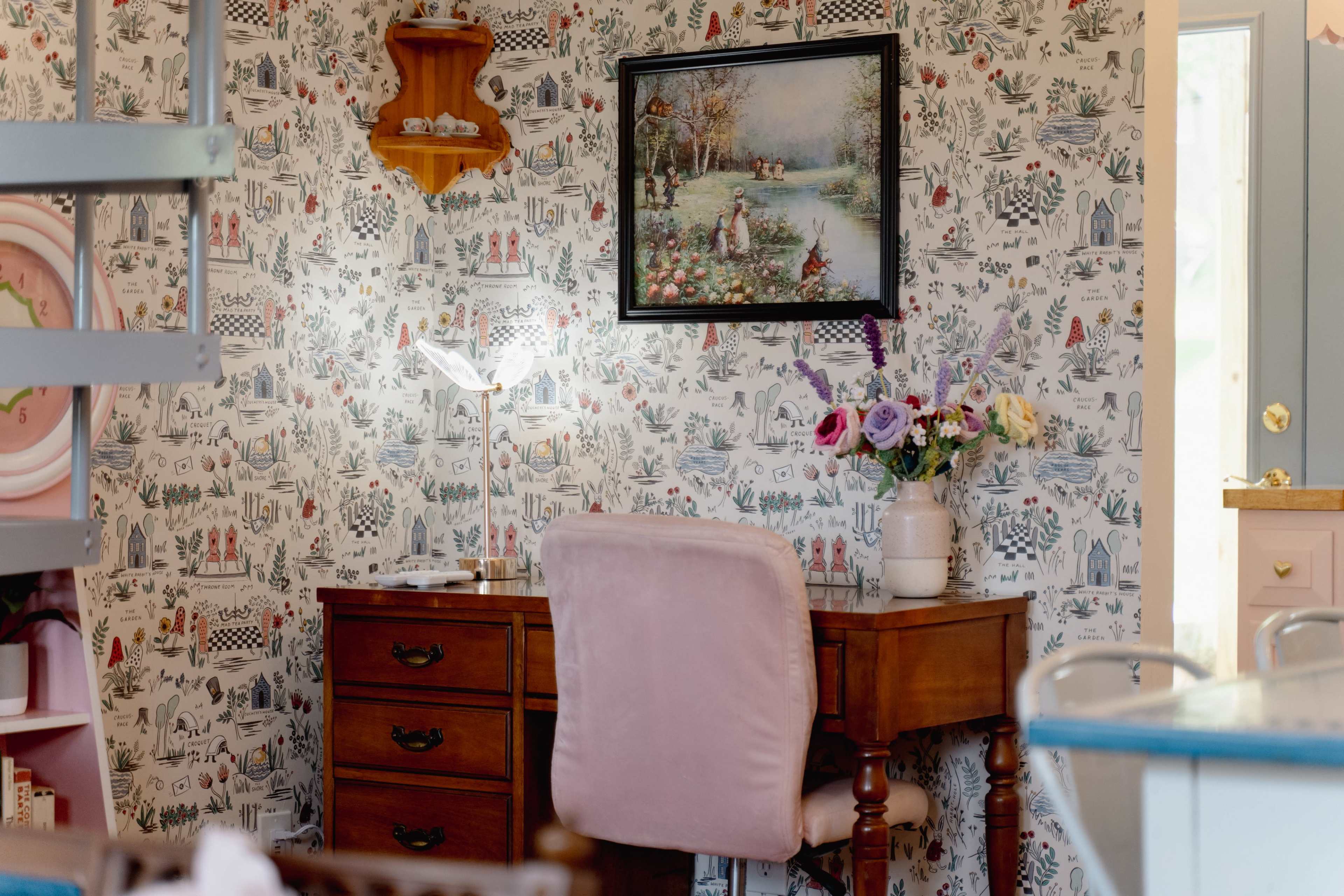 The scene features a wooden desk with a pink upholstered chair, adorned with a vase of flowers, set against a patterned wallpaper and a framed painting.