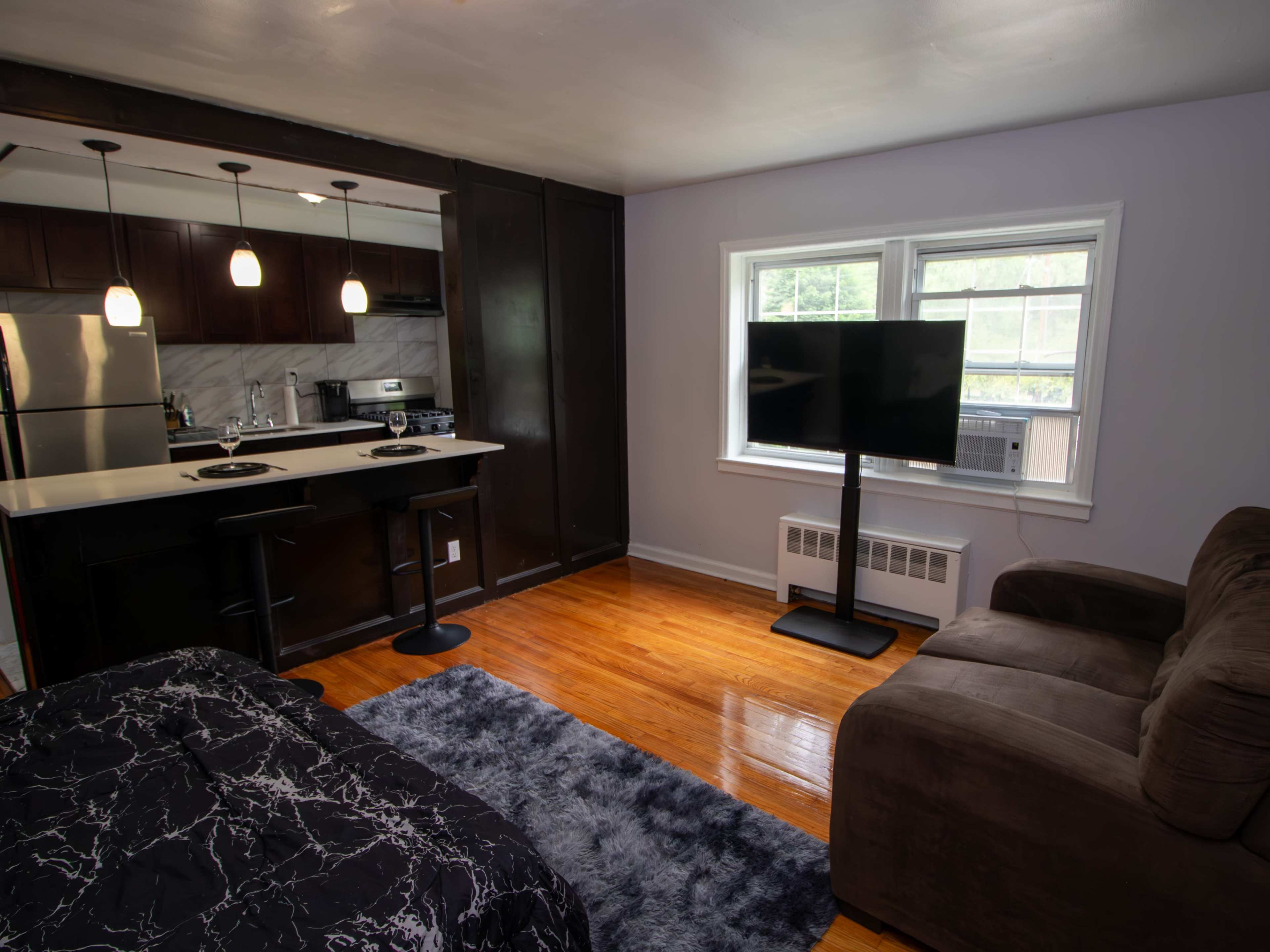The image shows a small living space with a kitchen area on one side, a couch, a TV on a stand, and a bed with a patterned coverlet.
