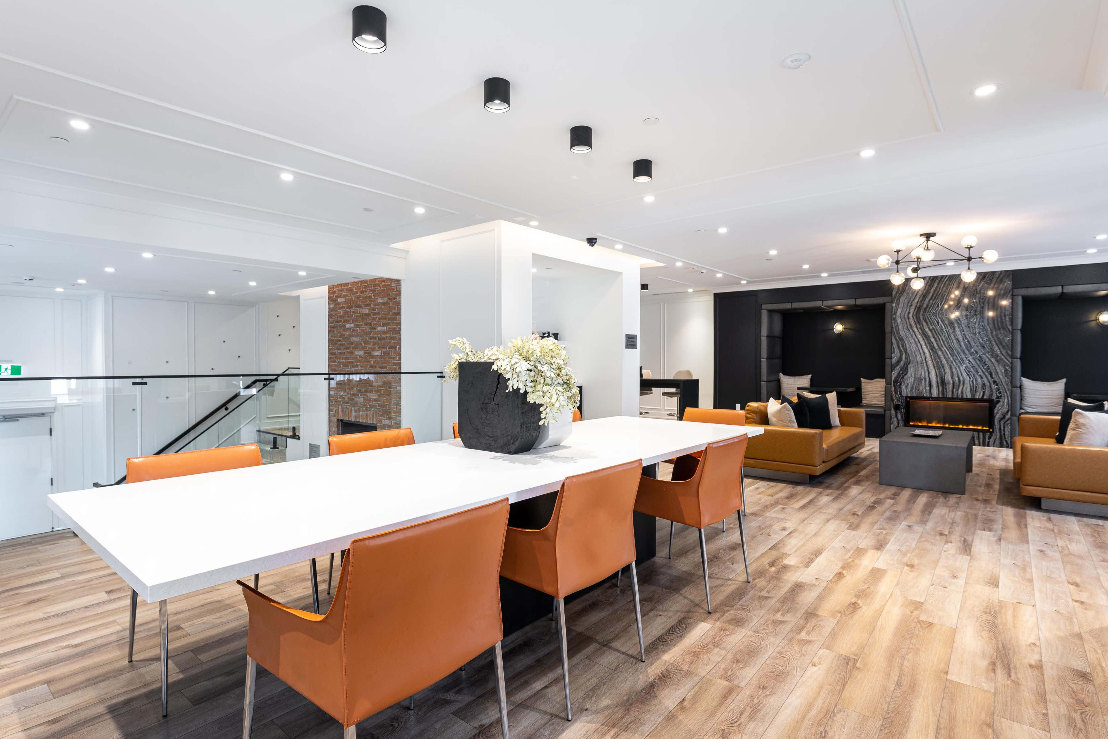 A modern dining area features a long white table surrounded by orange chairs, with a living space and fireplace visible in the background.