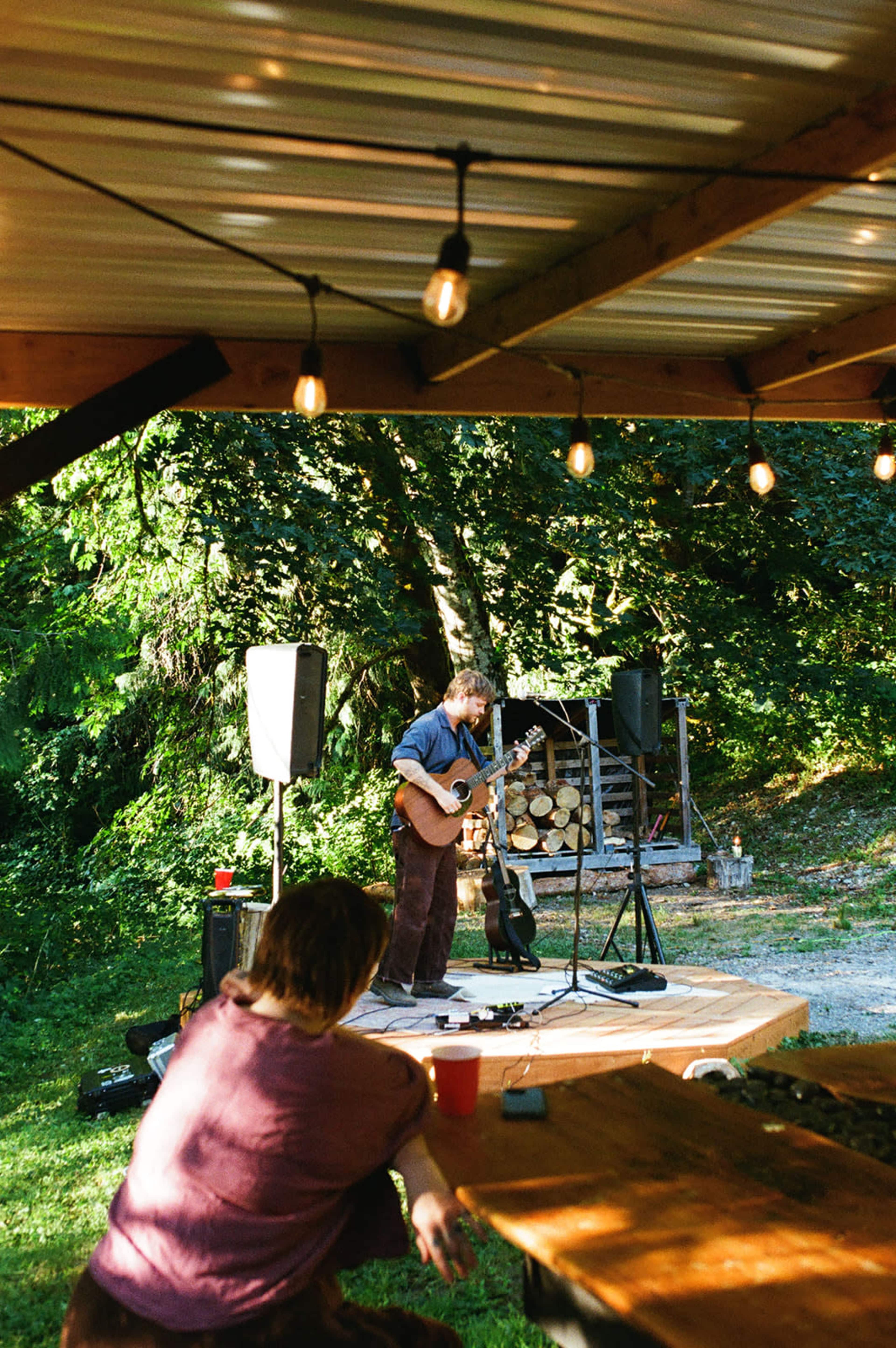 A musician plays guitar on a small outdoor stage surrounded by greenery, while a person sits at a nearby table watching.