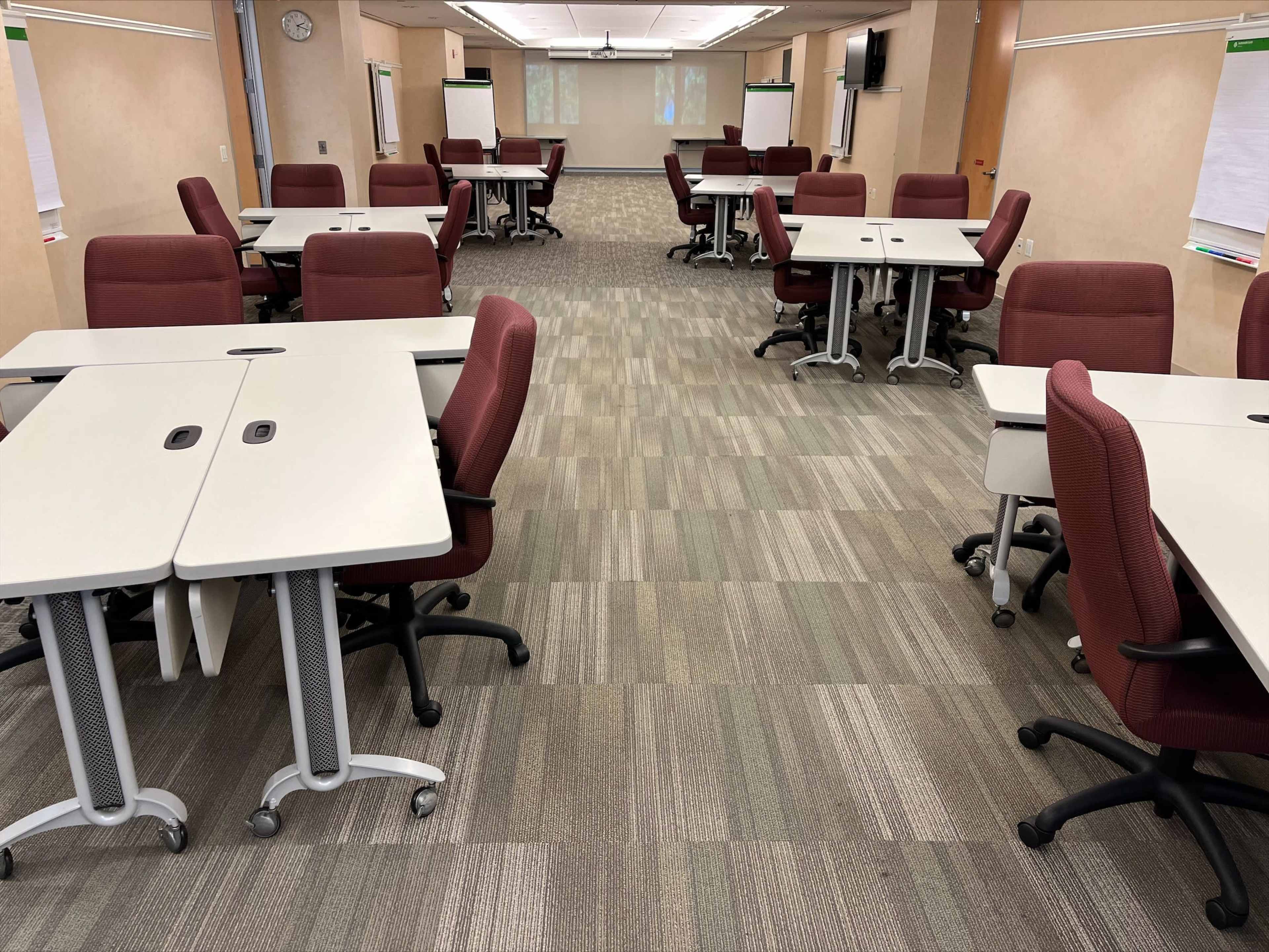 A rectangular classroom features multiple tables and chairs arranged in rows, with a neutral-colored carpet and beige walls.