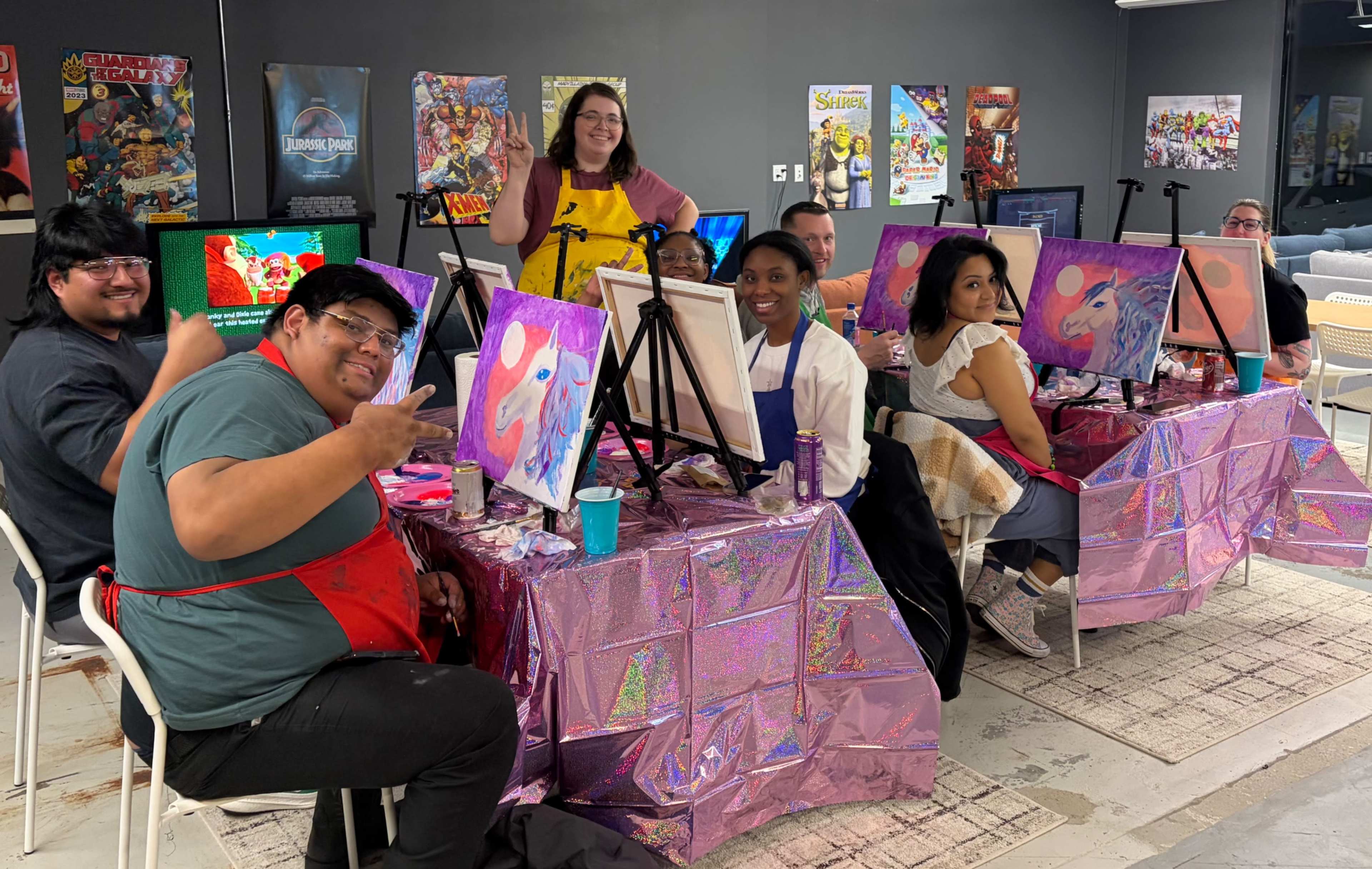A group of people sits at tables covered in shiny pink cloth, working on their paintings in a lively art studio.