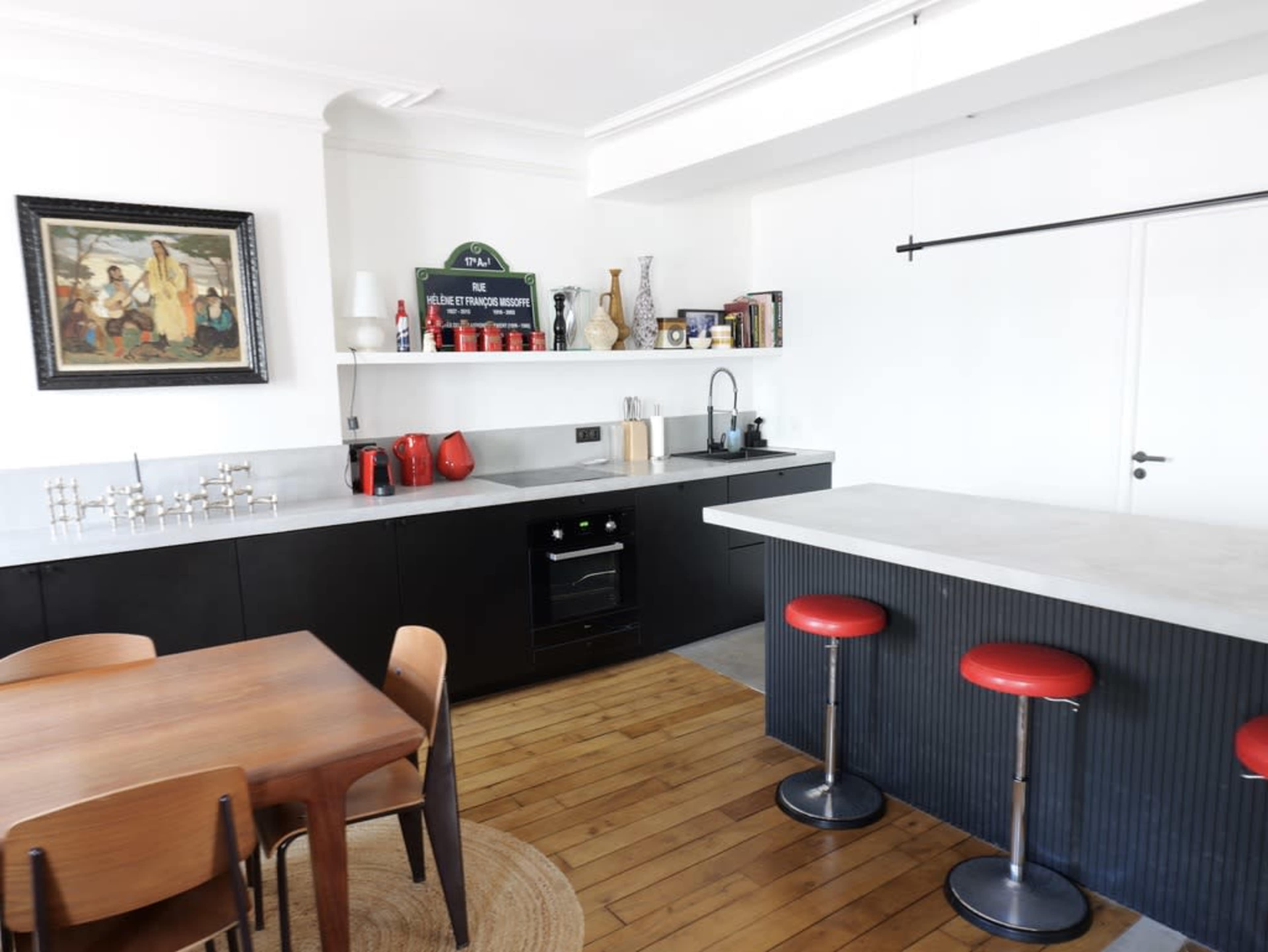 The image shows a modern kitchen with black cabinetry, a concrete countertop, red bar stools, and a wooden dining table, complemented by a framed painting on the wall.
