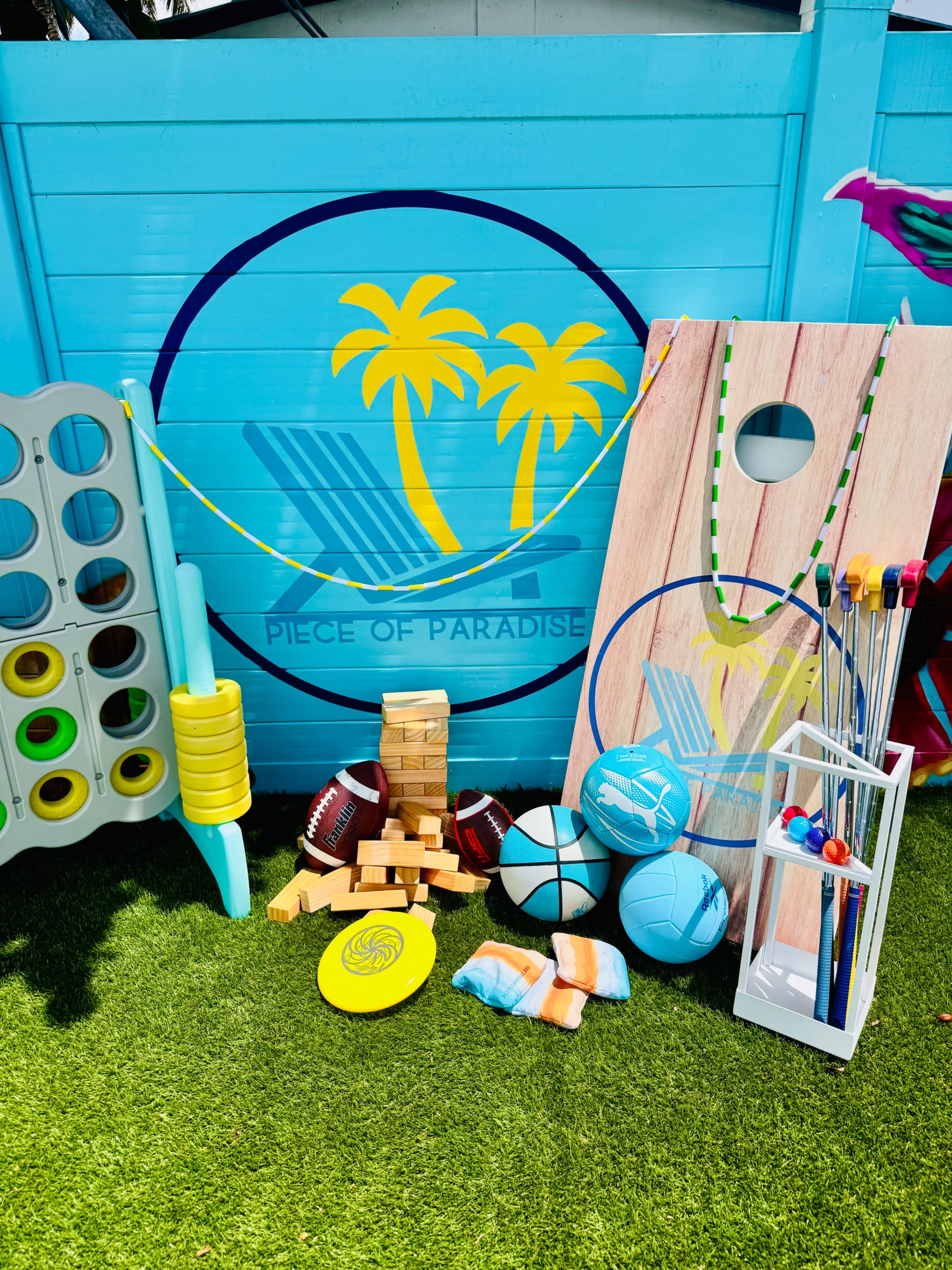 The image shows a colorful outdoor game area featuring various sports equipment, including balls, a frisbee, a stack of Jenga blocks, and a wooden board with a hole, all arranged against a blue wall decorated with palm trees.