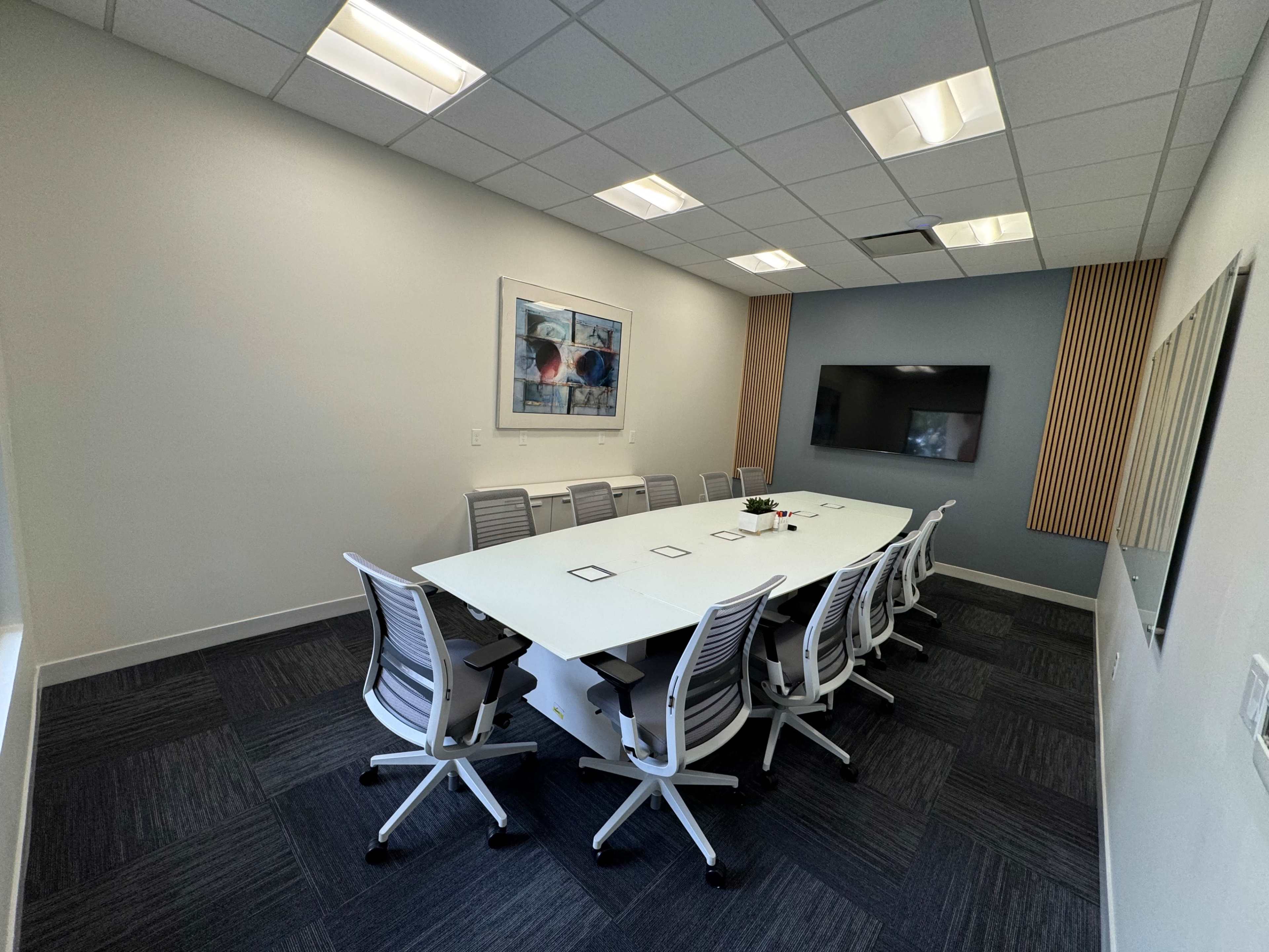 A bright conference room features a large white table surrounded by several gray chairs, with a wall-mounted screen and modern artwork.