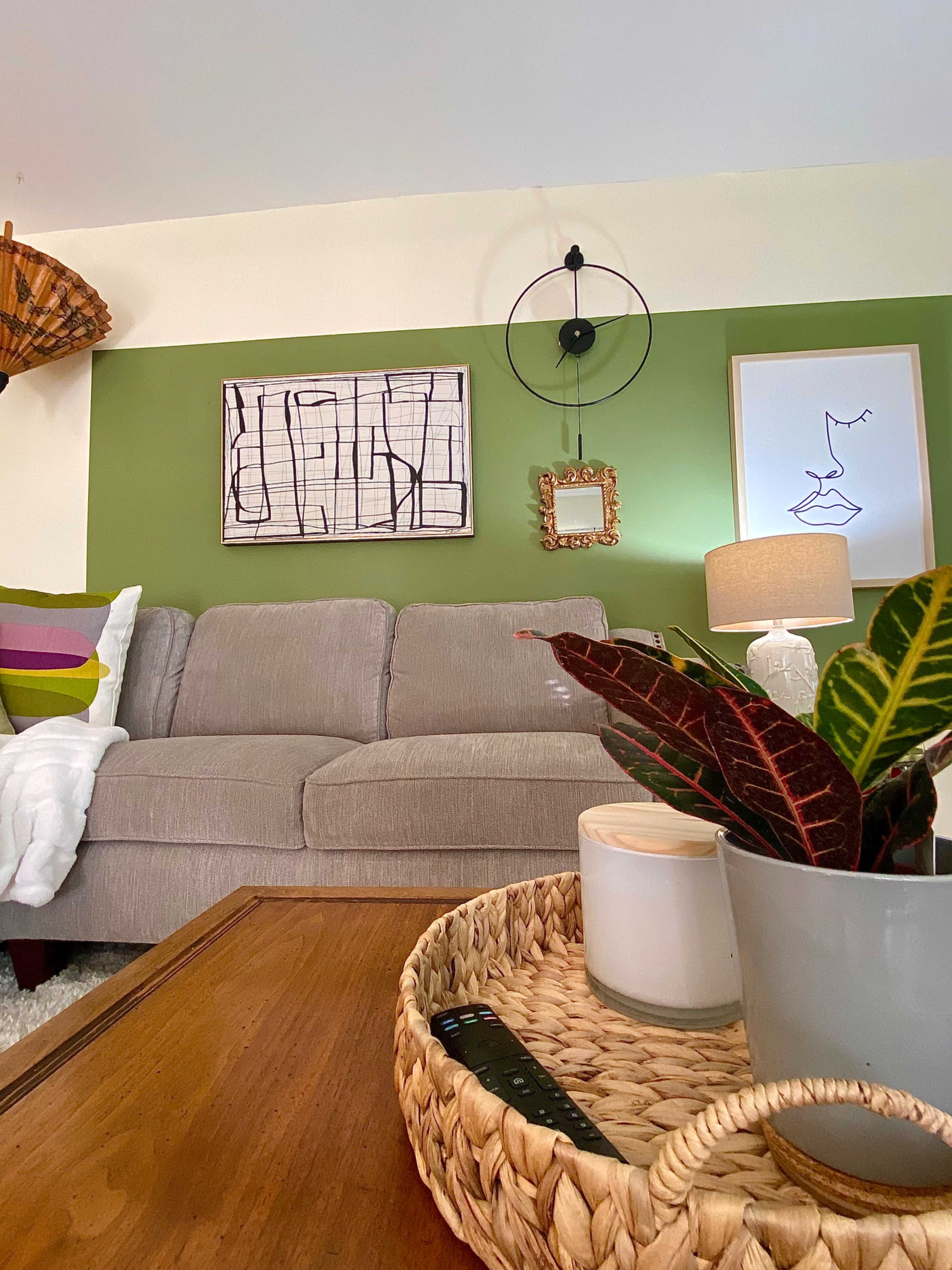 A gray couch is positioned in a living room with green walls, featuring a wooden coffee table with a woven tray, a potted plant, and framed artwork on the wall.