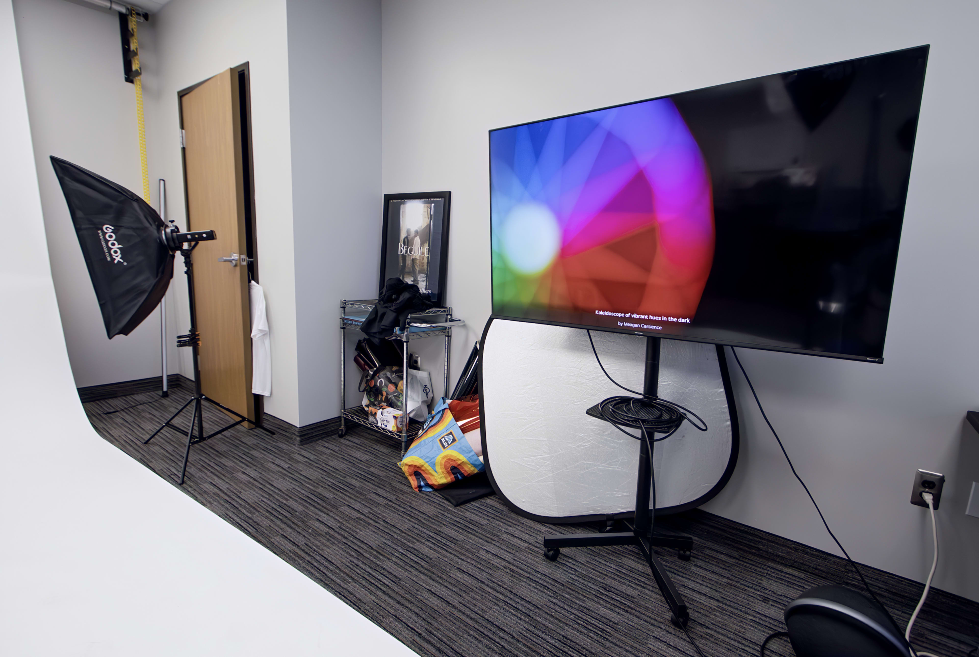 The image shows a studio setup featuring a large television on a stand, a light umbrella, and various items stored on a shelf in a plain room.