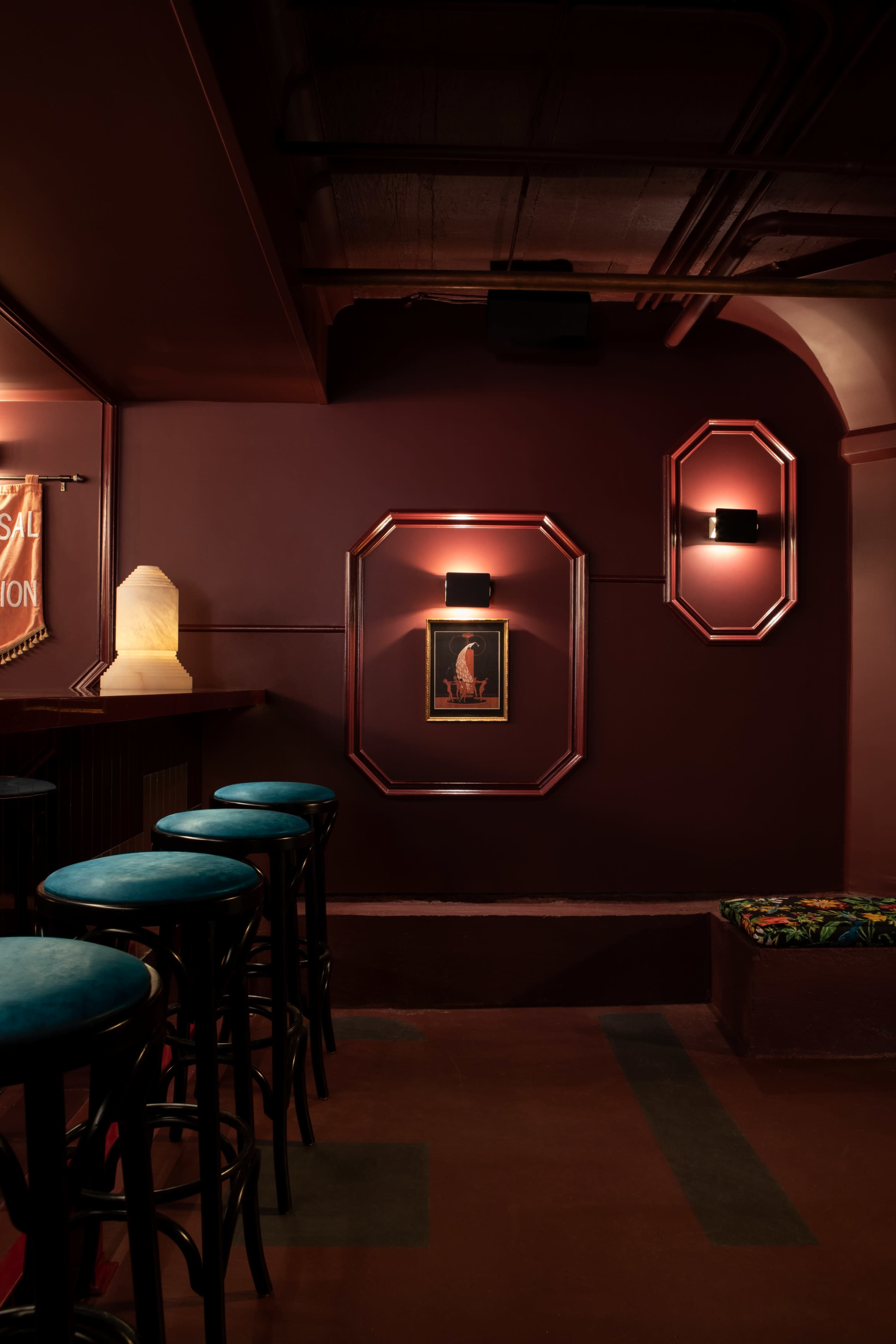 The image shows a dimly lit bar interior featuring dark red walls, illuminated wall art, and several bar stools.