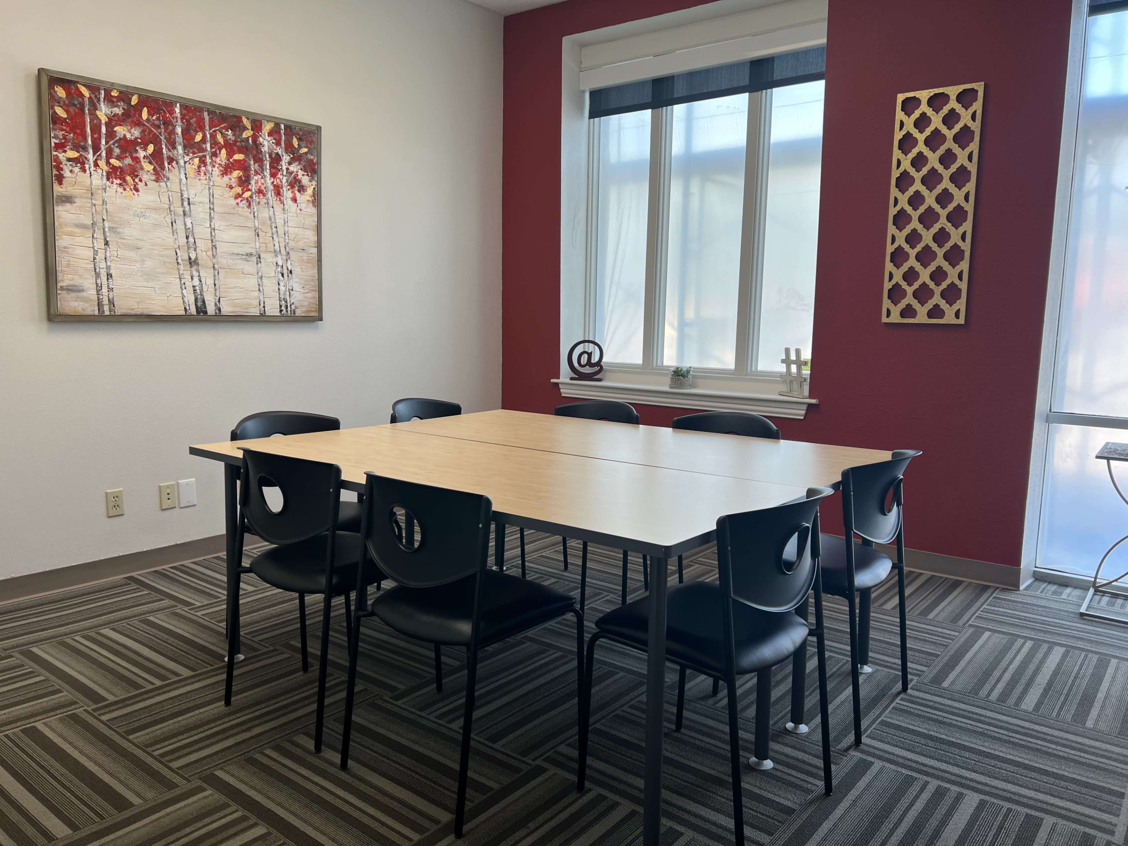 A conference room features a rectangular table surrounded by six black chairs, with a framed artwork on the wall and light streaming through a large window.
